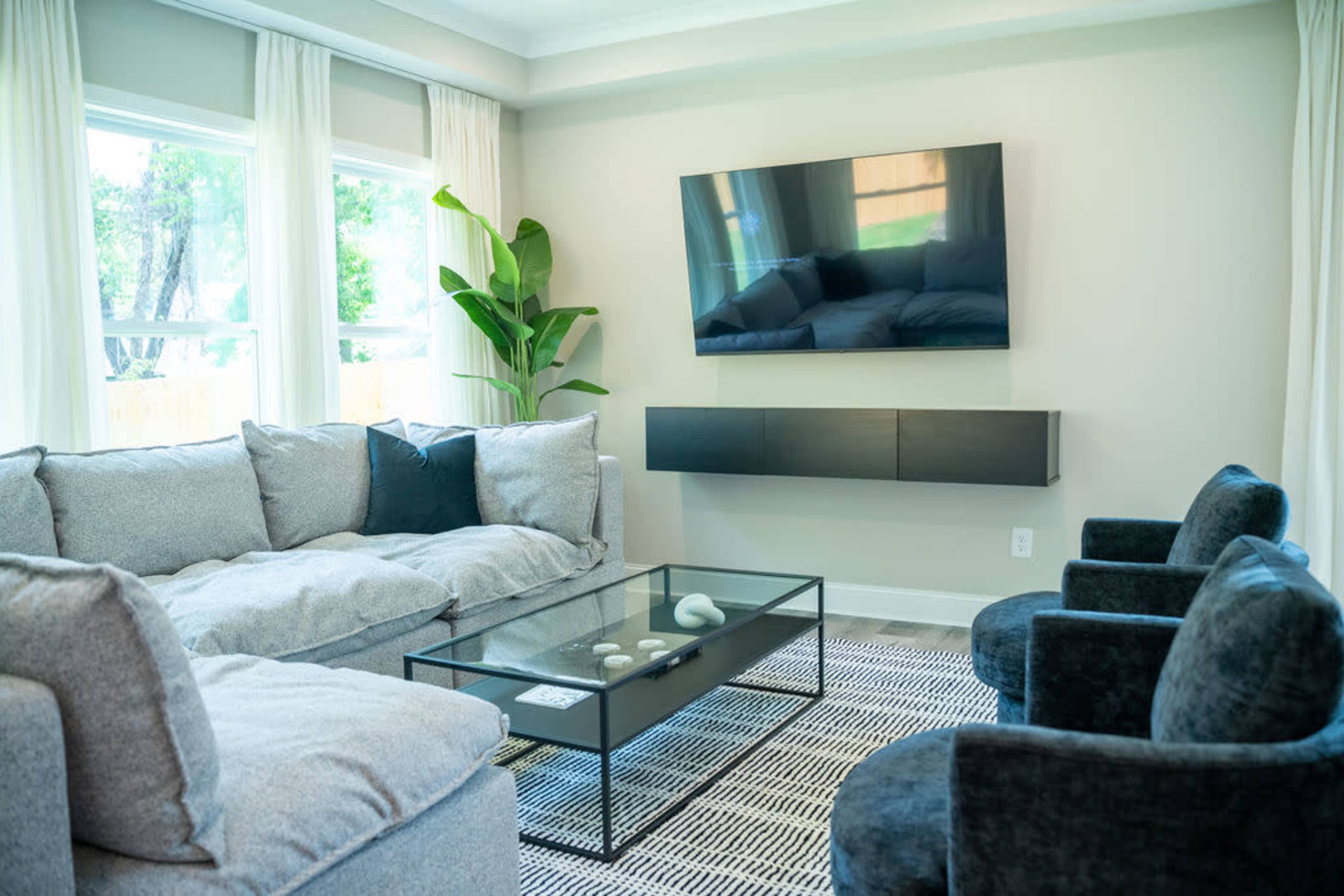 A modern living room featuring a grey sectional sofa, two black chairs, a glass coffee table, and a wall-mounted television.