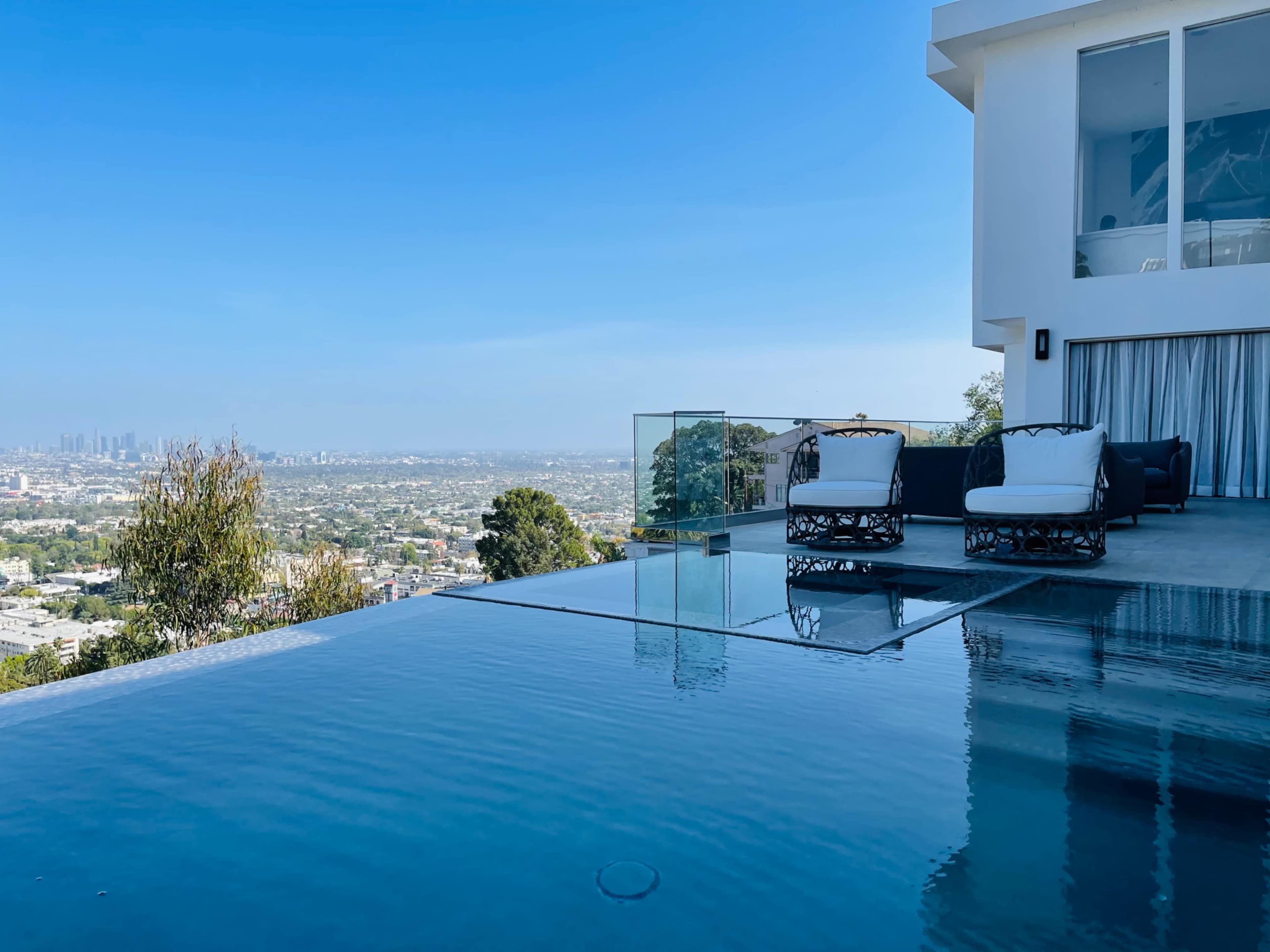 The image shows a modern hillside villa with an infinity pool overlooking a city skyline under a clear blue sky.