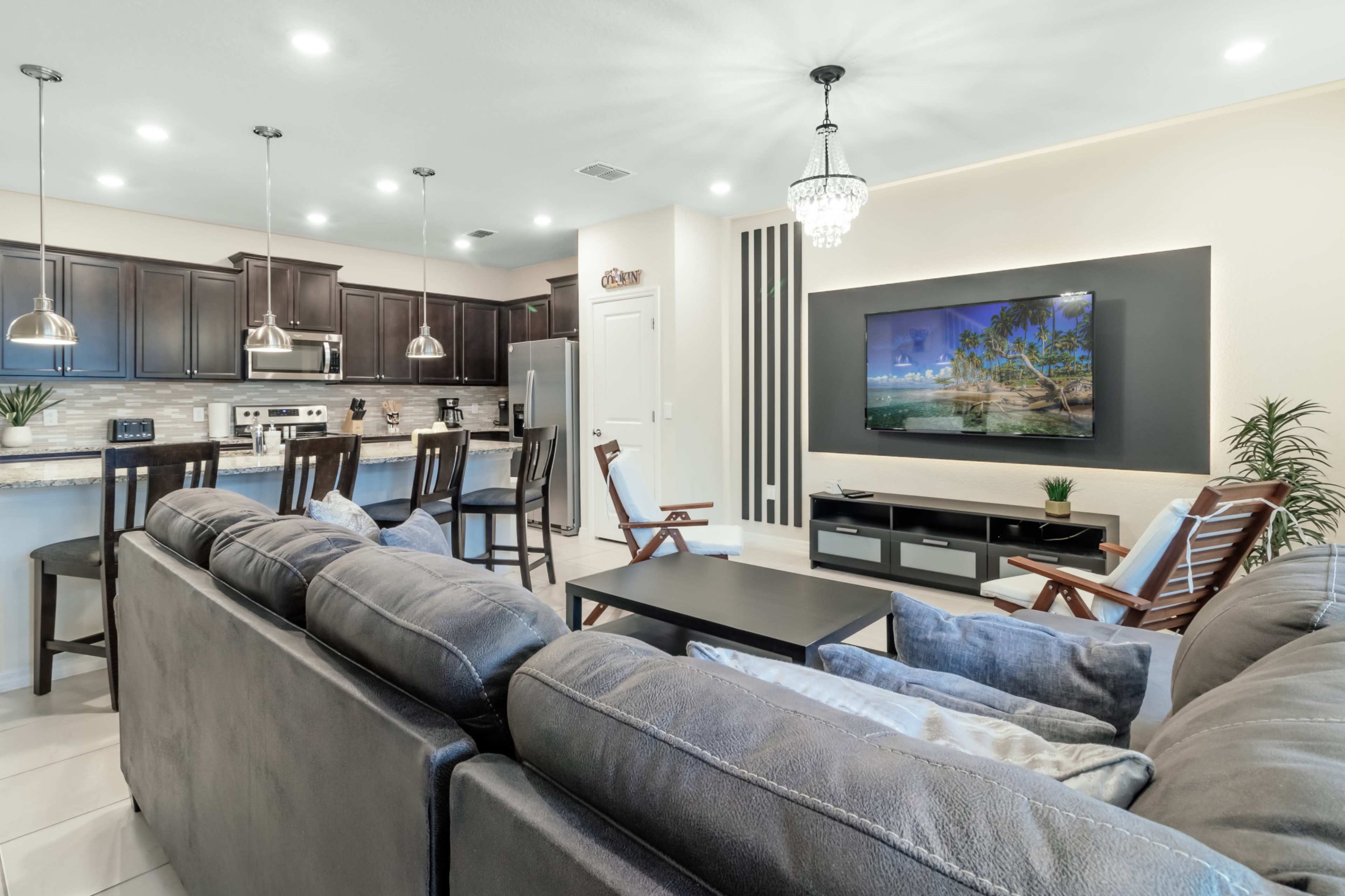 A modern living area with a gray sectional sofa, a coffee table, and a wall-mounted television, adjacent to a kitchen with dark cabinetry and stainless steel appliances.