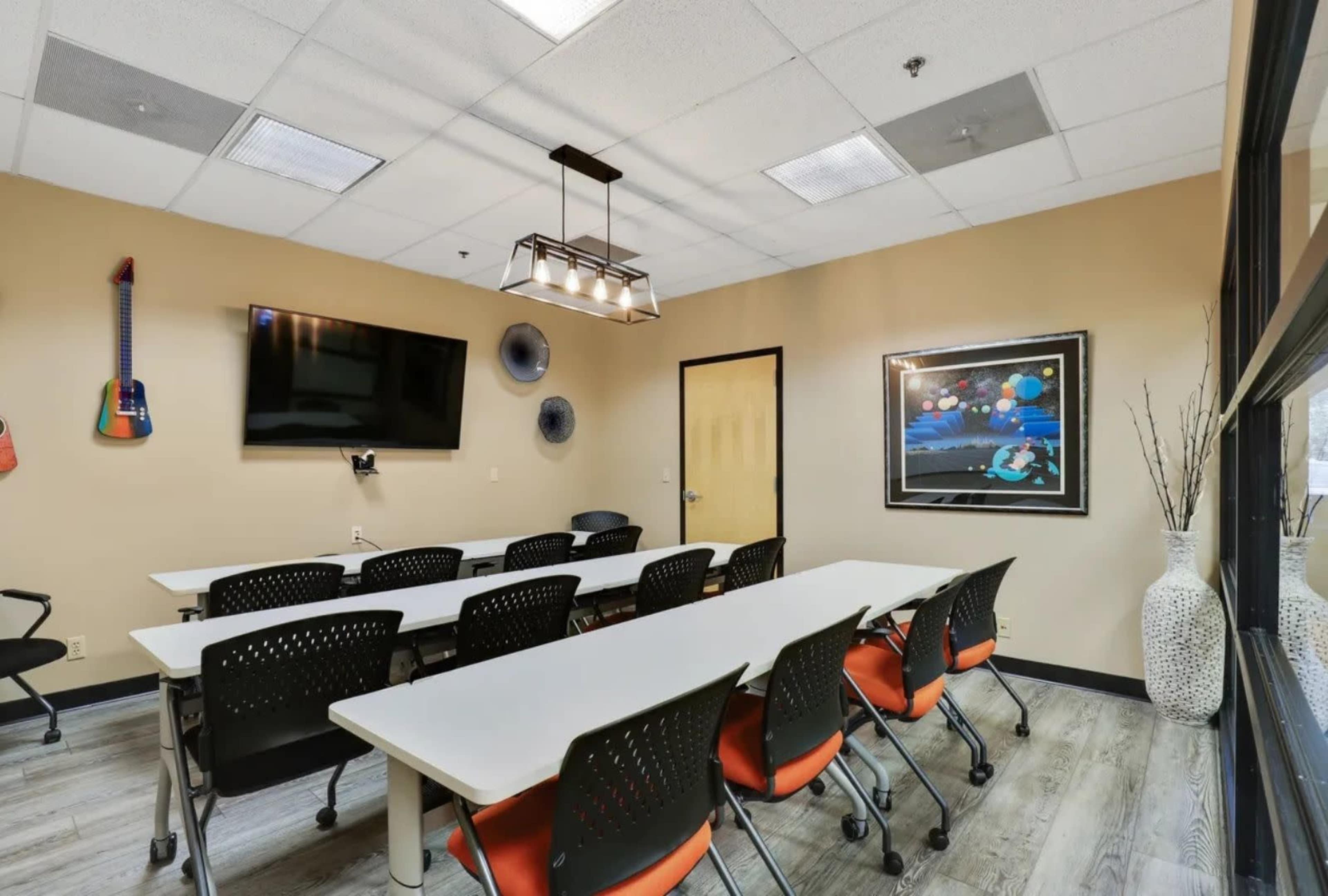The image shows a conference room with a long white table and black chairs, equipped with a television and wall art.