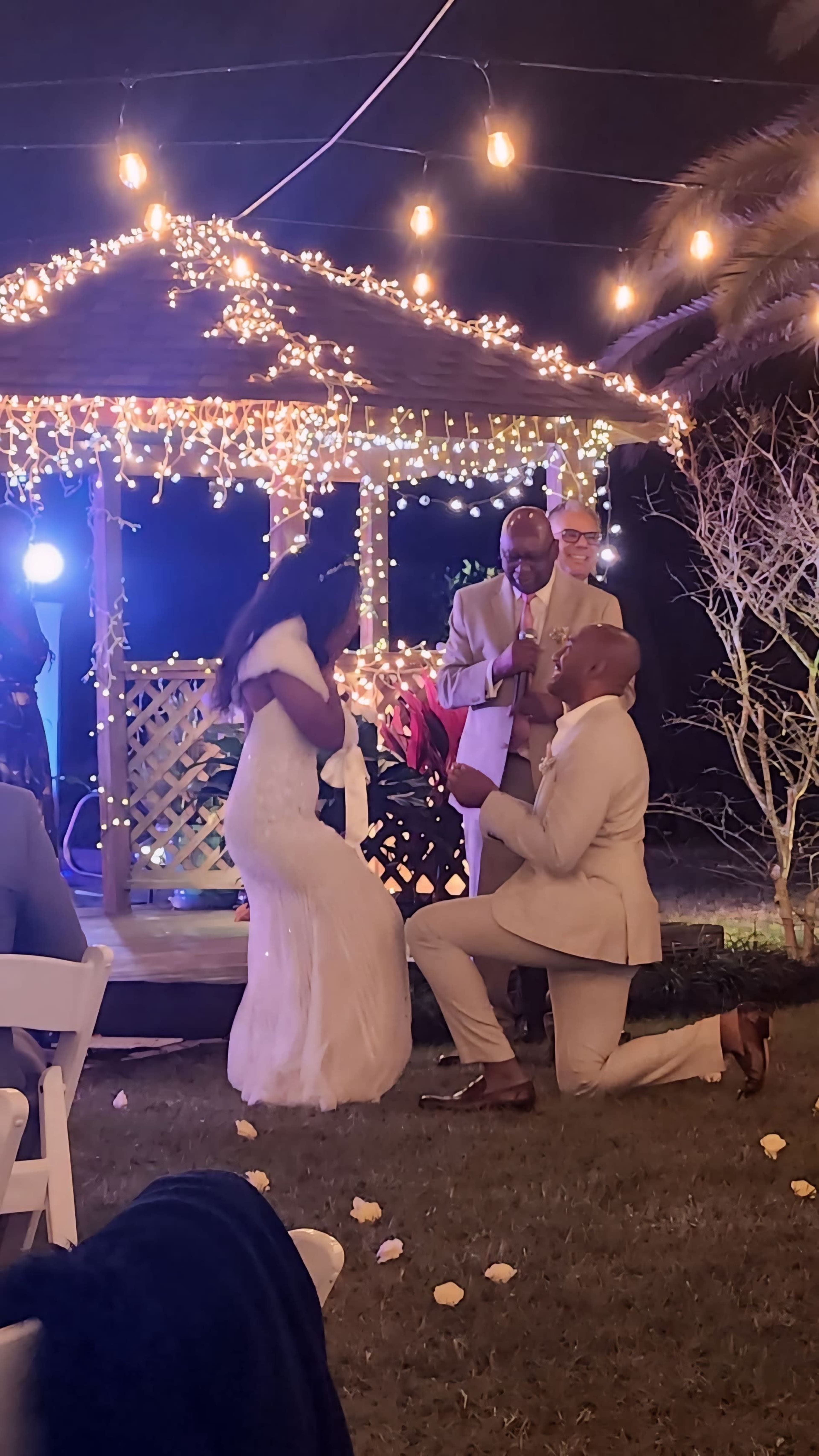 A man kneels with a ring in front of a woman in a wedding dress as a group celebrates under a decorated gazebo lit with string lights.