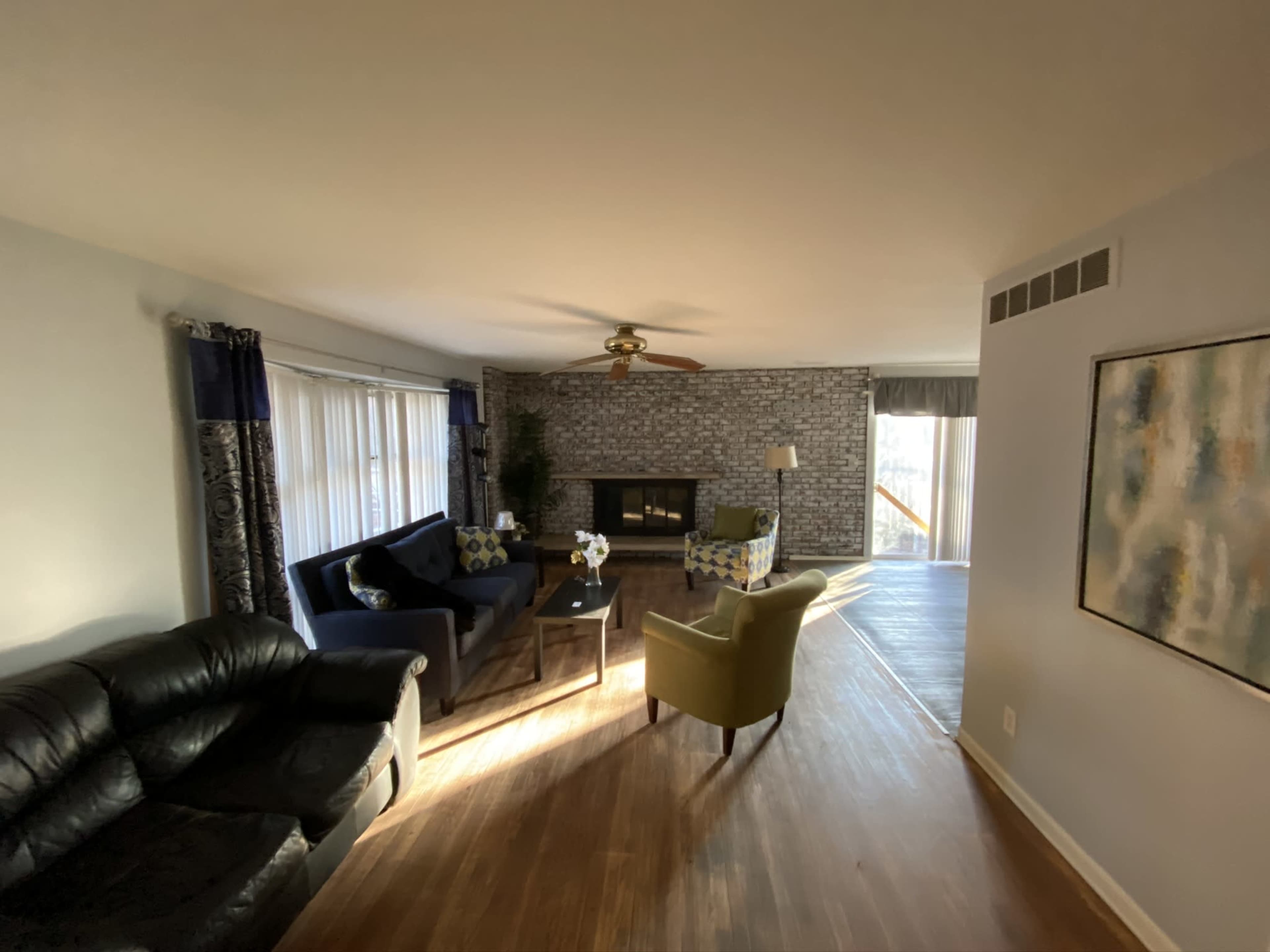 A living room with a black leather sofa, a blue couch with decorative pillows, a coffee table, and a fireplace, illuminated by natural light from large windows.