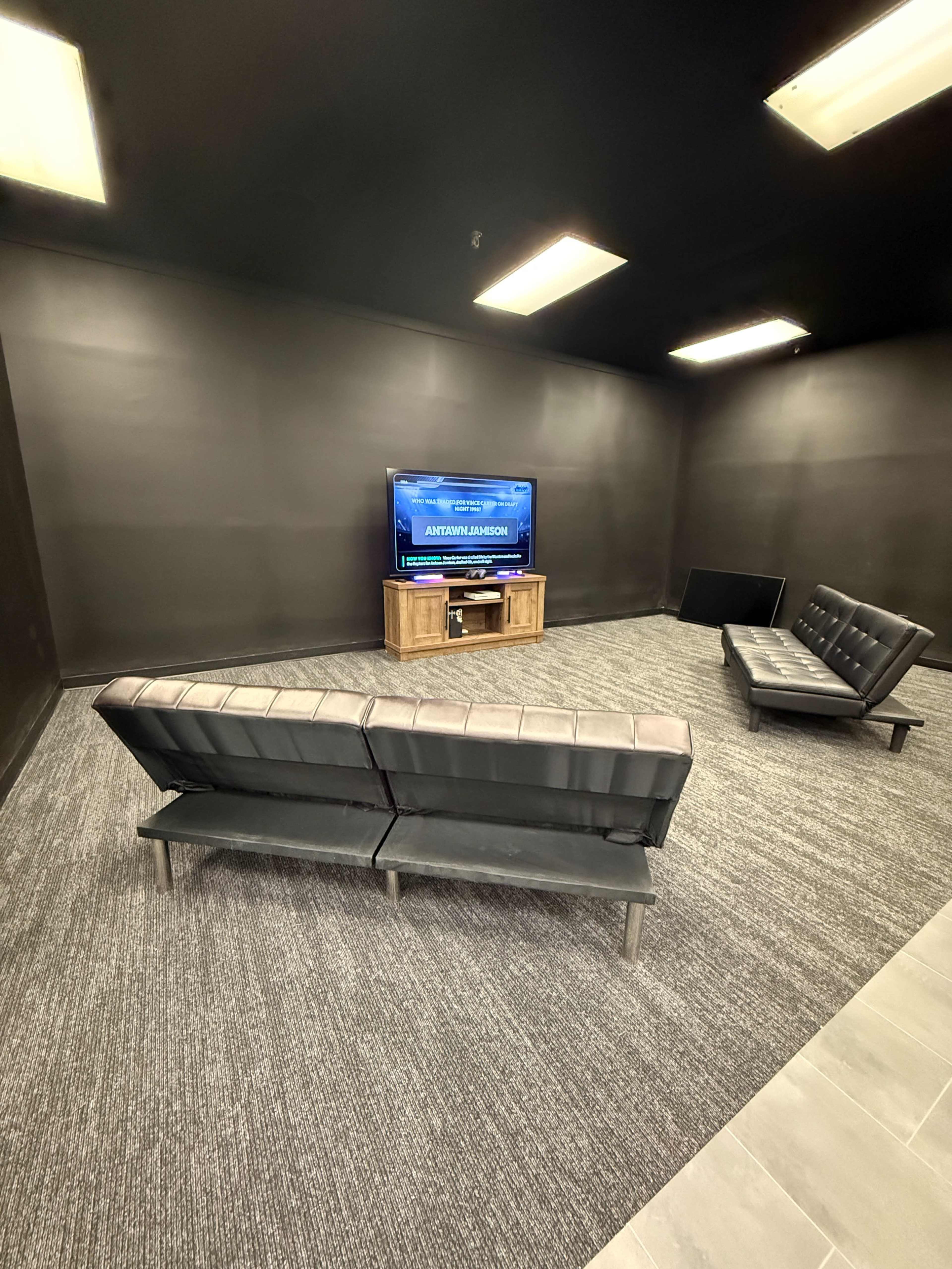 The image shows a sparsely furnished room with black walls, featuring a television on a wooden stand, a brown leather couch, and two additional seating options against the walls.