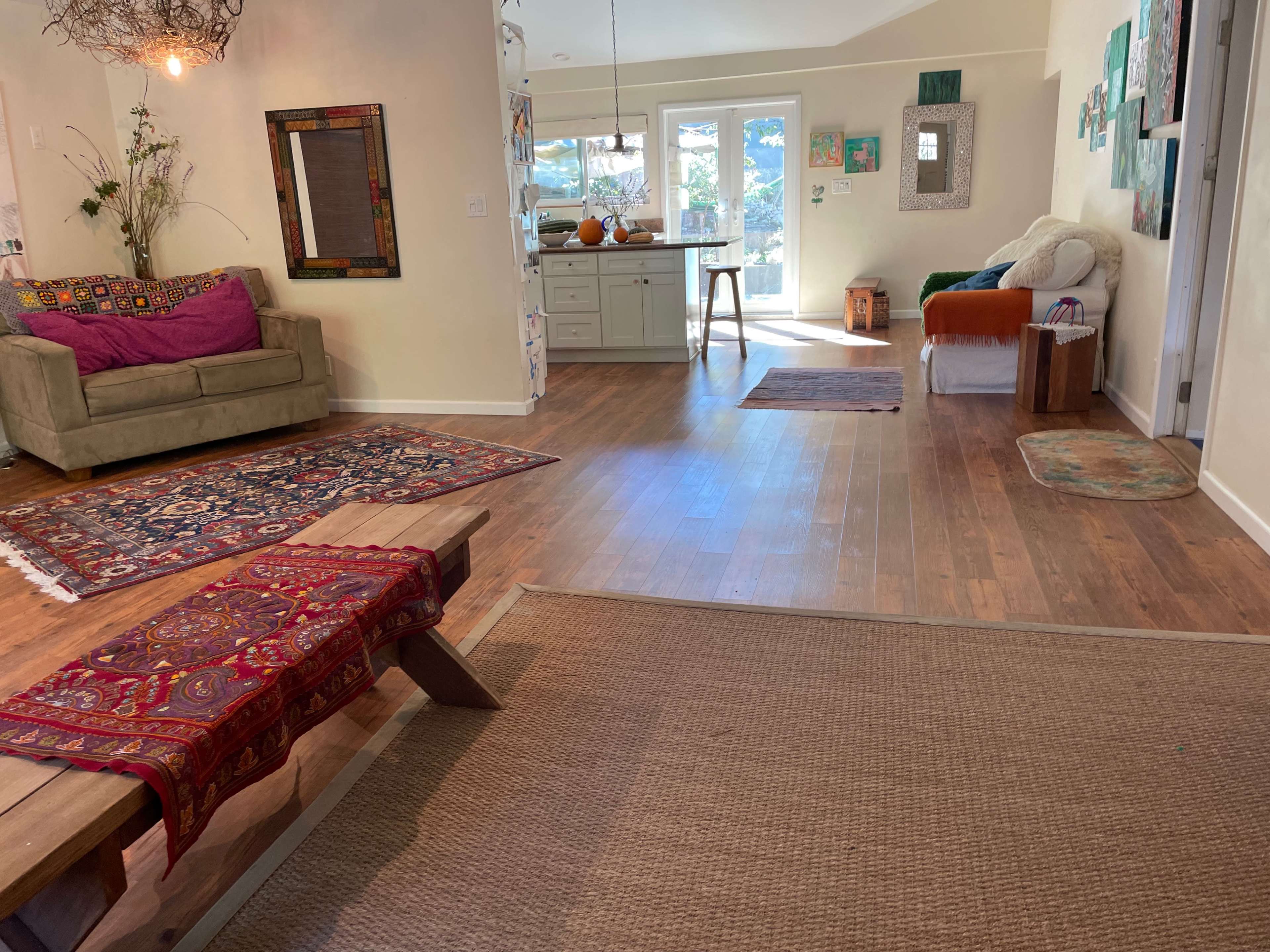A spacious living area featuring a green sofa, decorative rugs, and an open kitchen area with sunlight streaming through large windows.