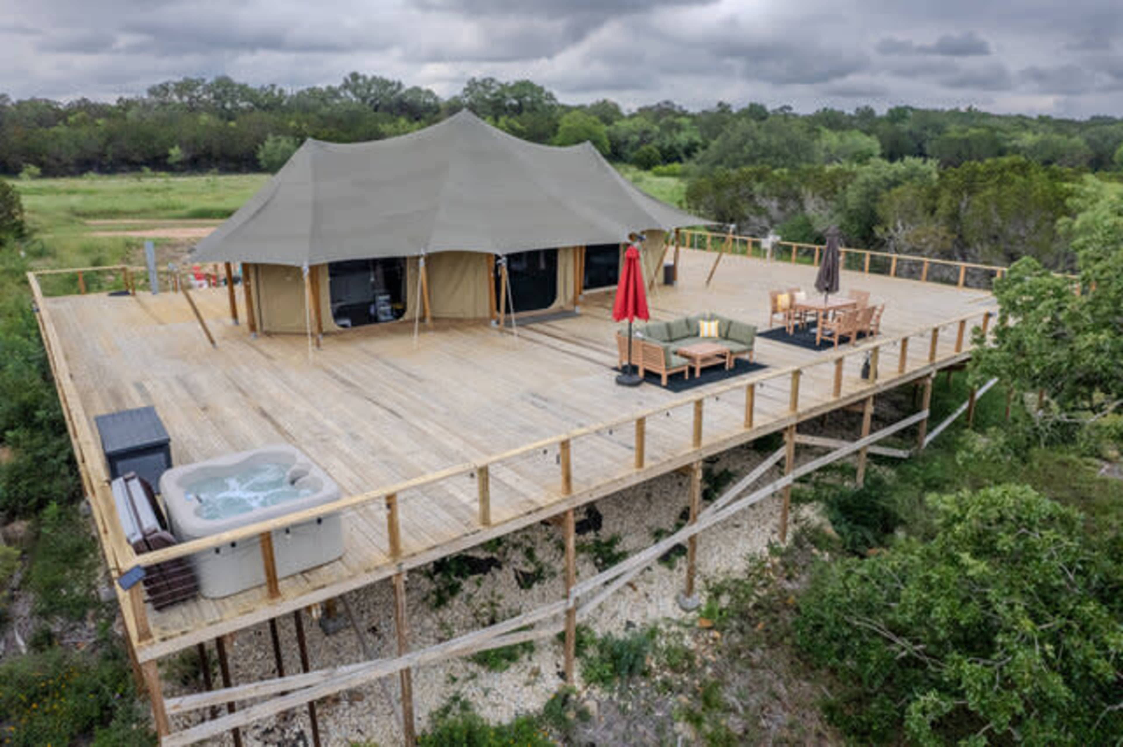 A large tent structure is raised on a wooden deck overlooking a green landscape, featuring a hot tub and outdoor seating areas.