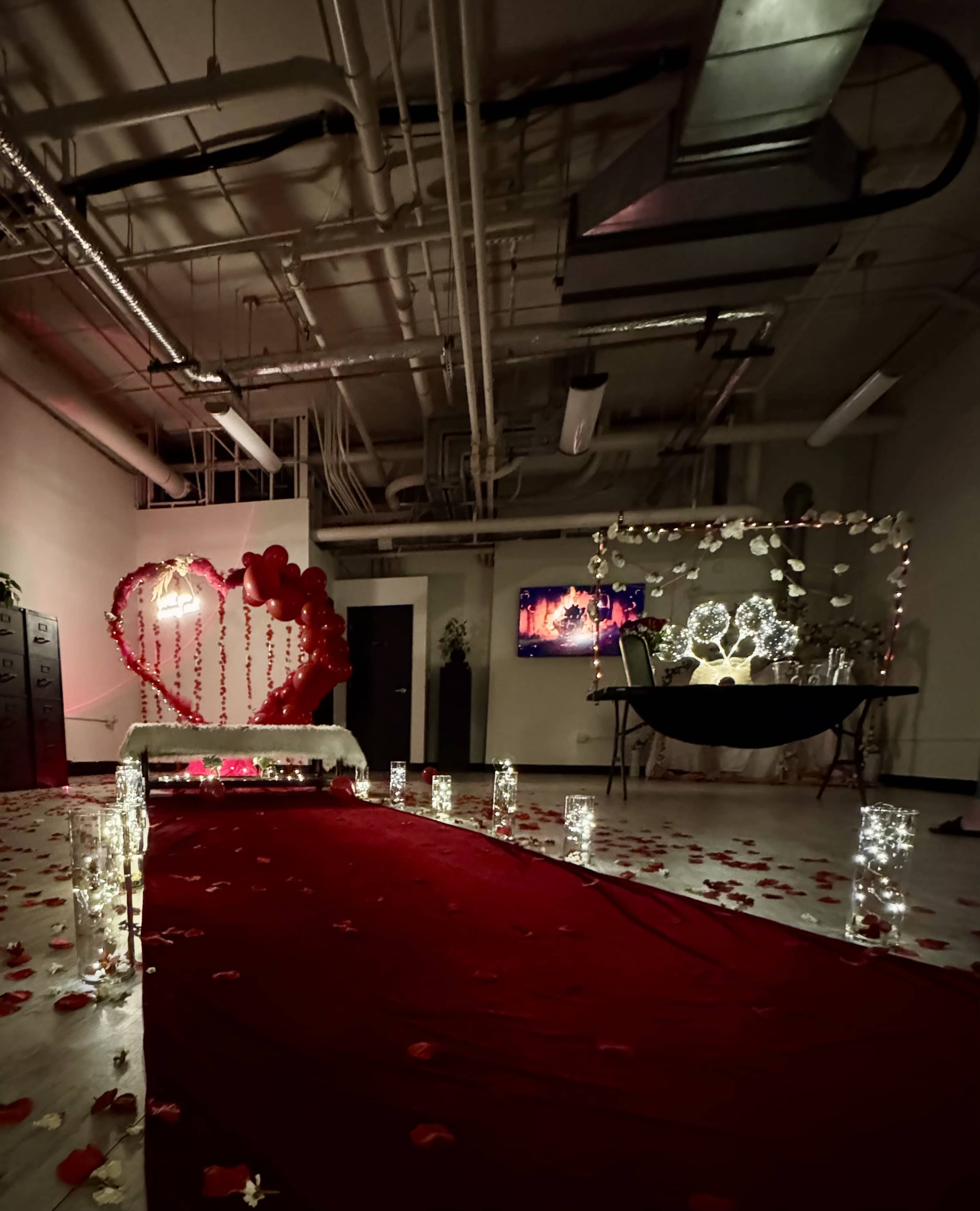 The image depicts a romantic themed room with a red carpet leading to a decorated table, surrounded by rose petals, candles, and heart-shaped decor, with a screen displaying a video in the background.