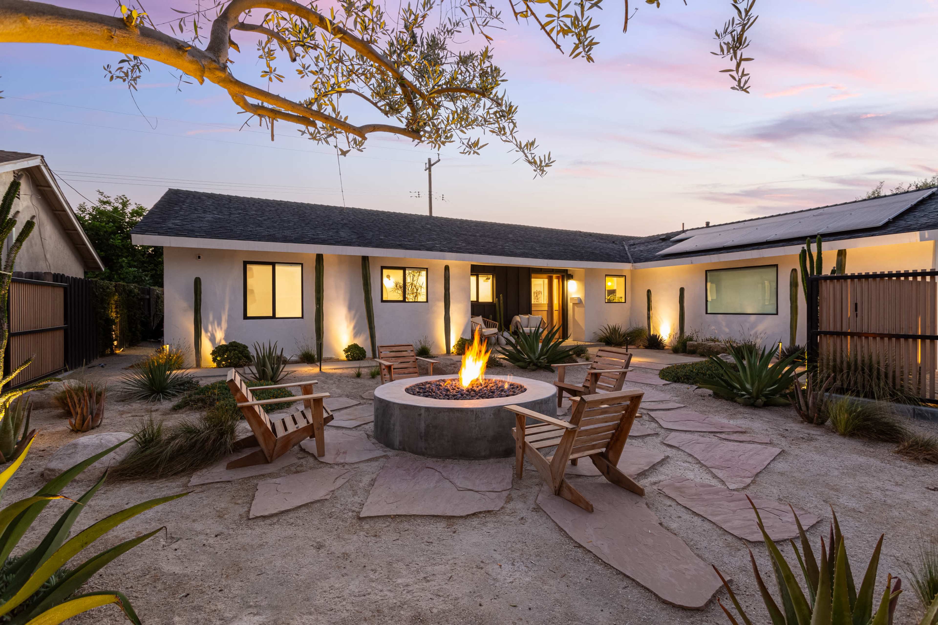 A circular fire pit surrounded by wooden chairs is set in a landscaped courtyard with desert plants and a single-story house in the background.