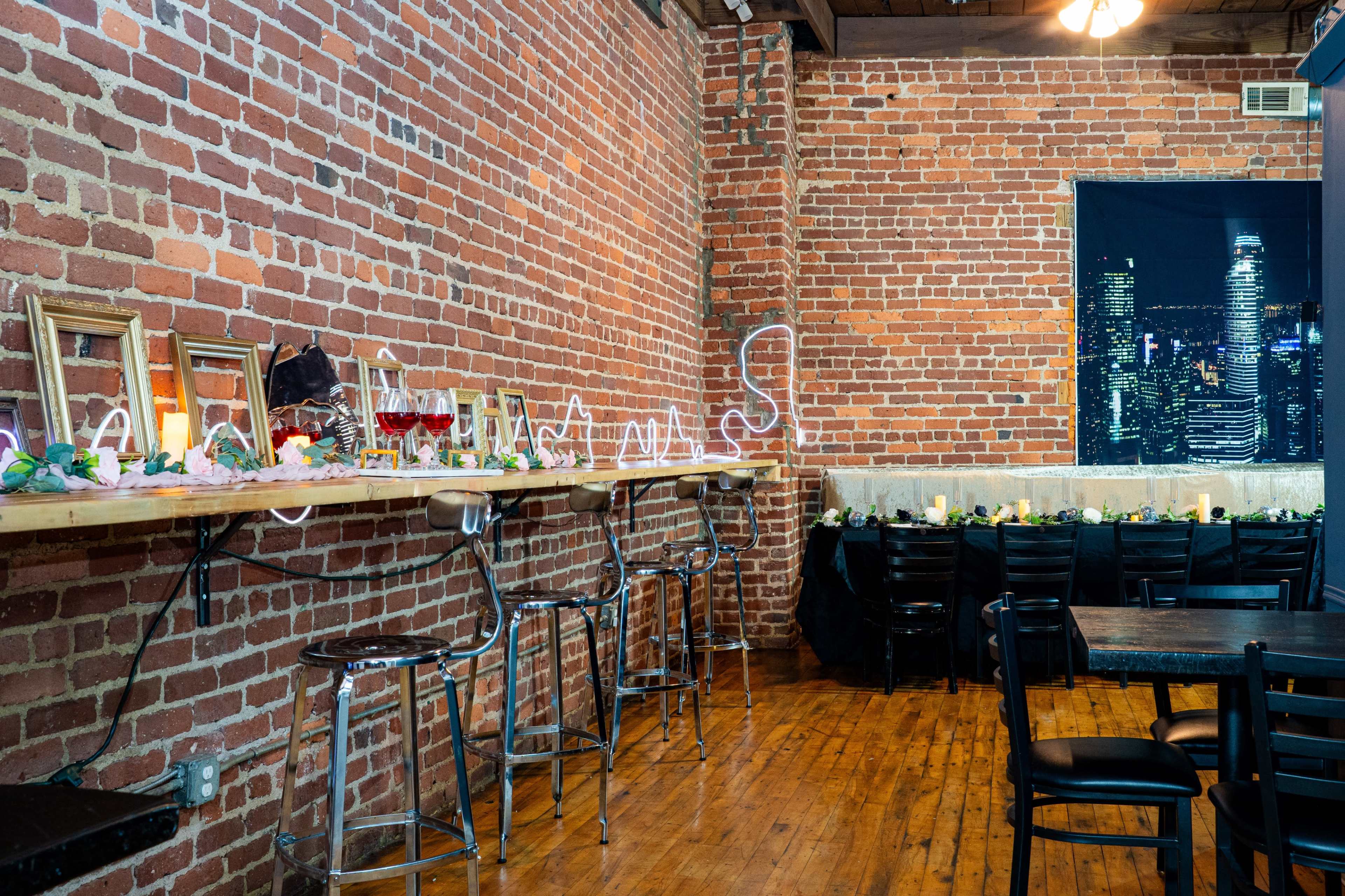 The image shows a rustic interior with exposed brick walls, a wooden bar lined with framed mirrors, and chairs, while a long table adorned with flowers and candles is situated in the background.
