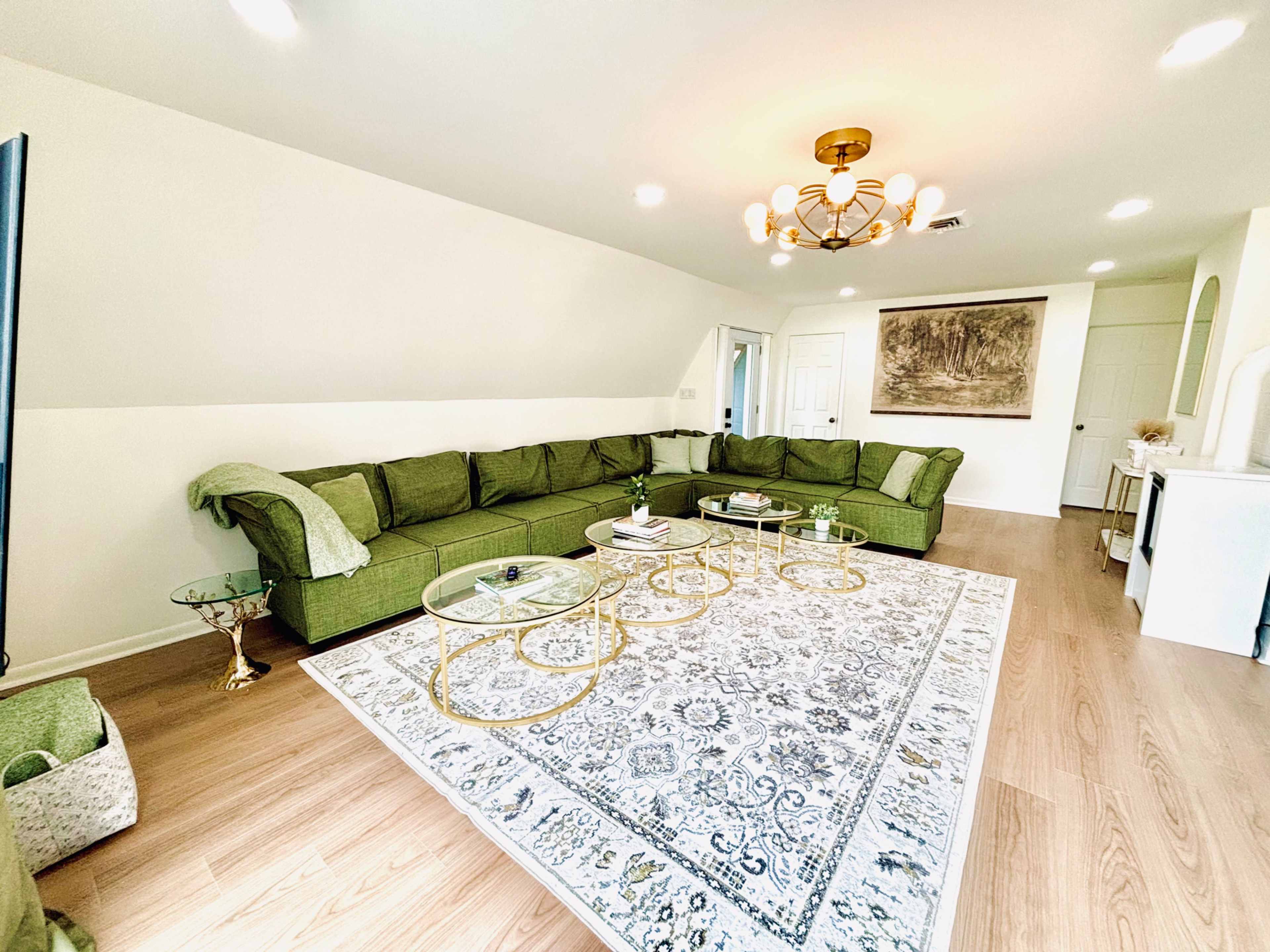 A modern living room with a large green sectional sofa, a patterned area rug, and a chandelier, accompanied by small gold coffee tables and decorative elements.