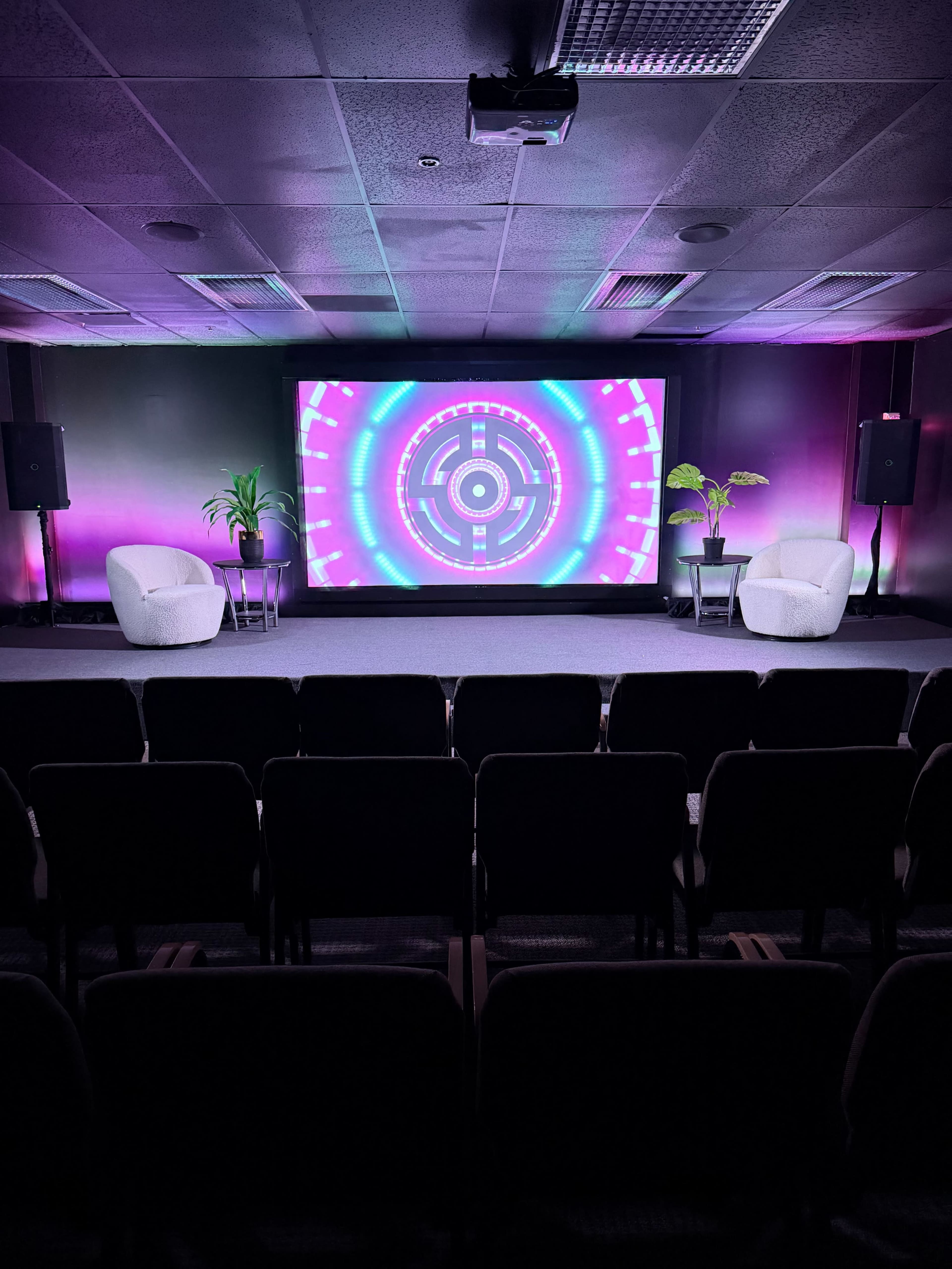 The image shows a stage setup with two white chairs, a large colorful screen displaying a circular design, and plants on either side, all positioned in front of rows of black seats.