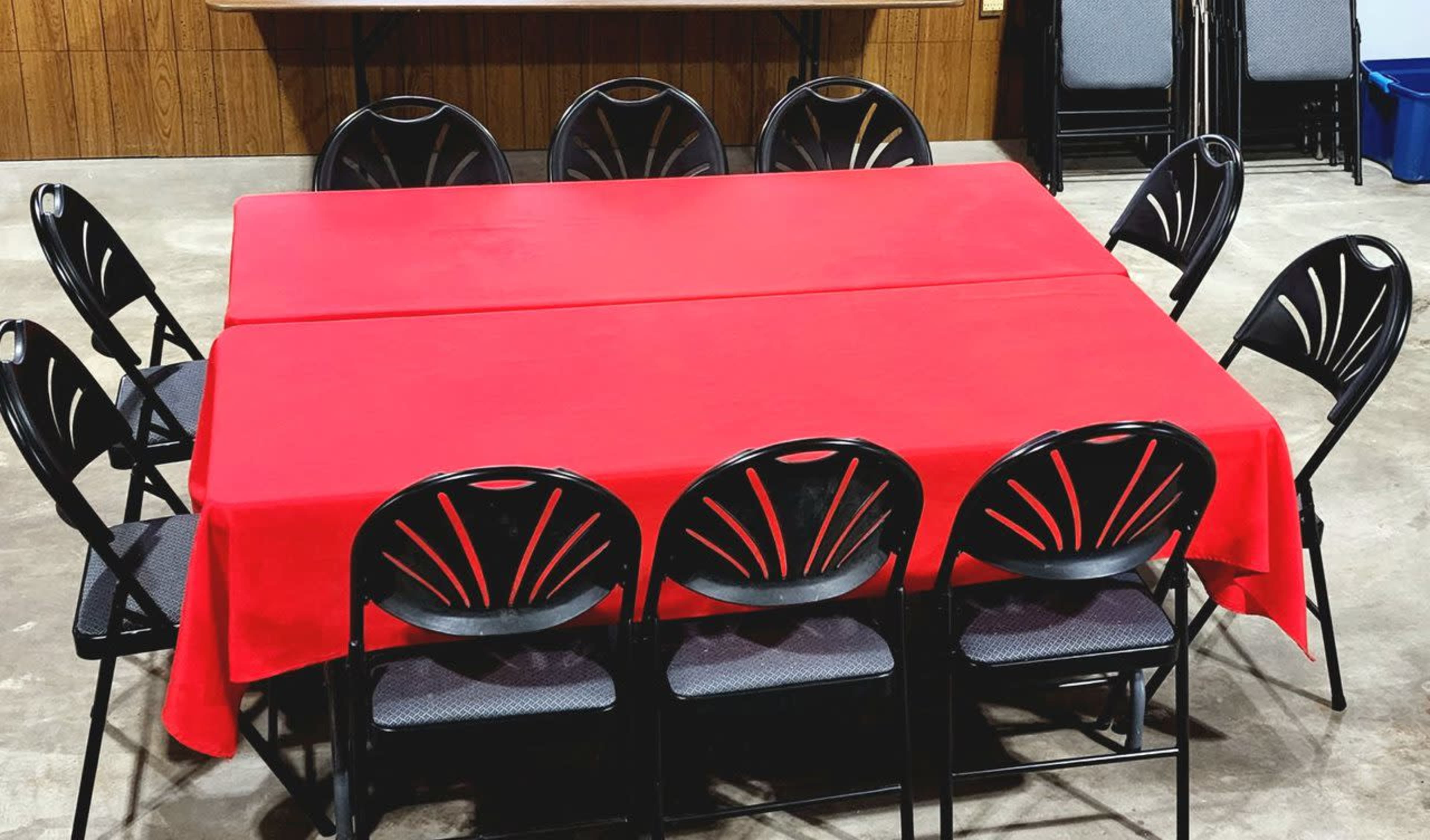 A square table covered with a red tablecloth is surrounded by ten black folding chairs in a room with wooden walls.