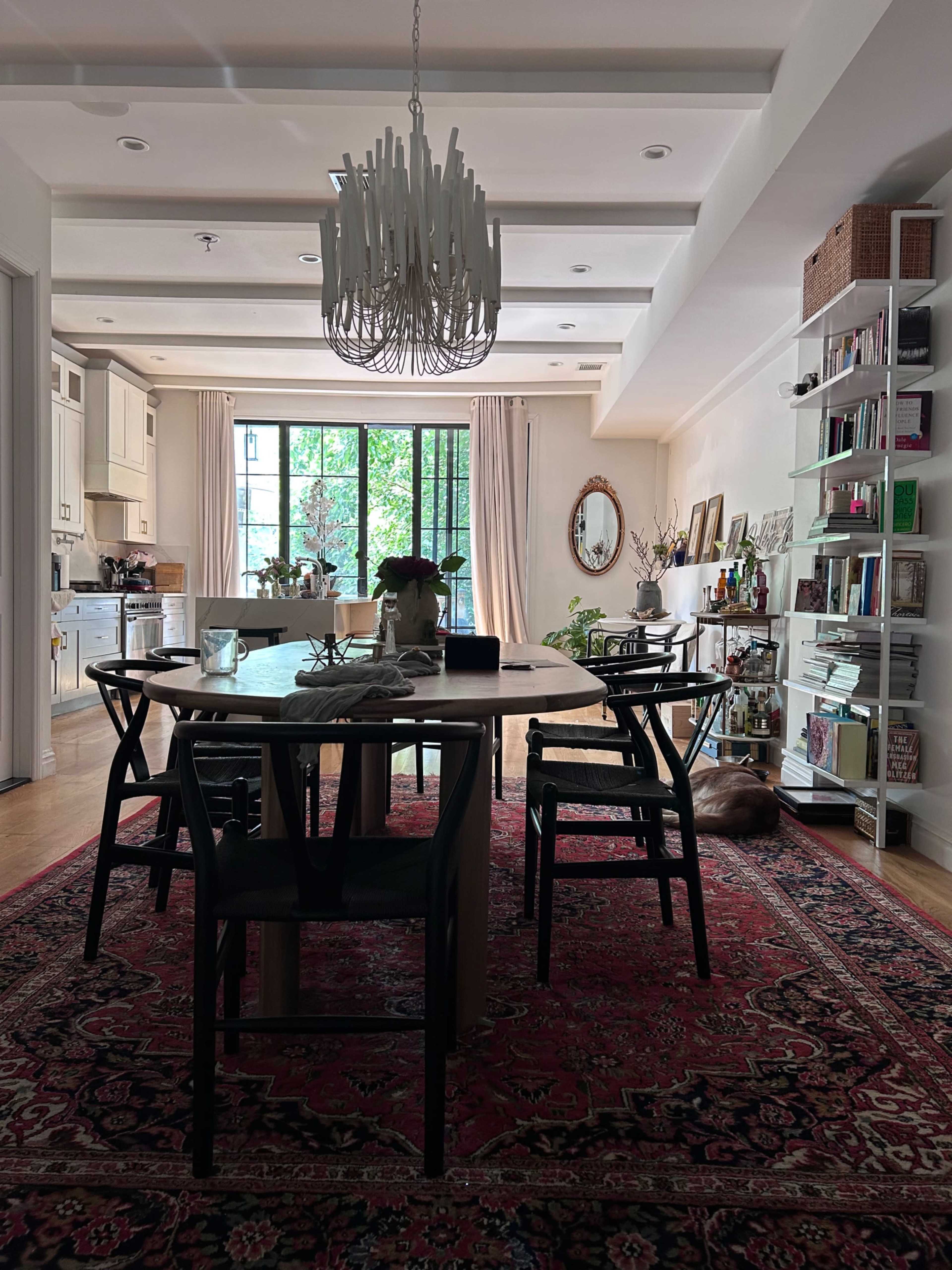 A dining area features a wooden table surrounded by black chairs, a large chandelier overhead, and shelves filled with books and decor against a wall.