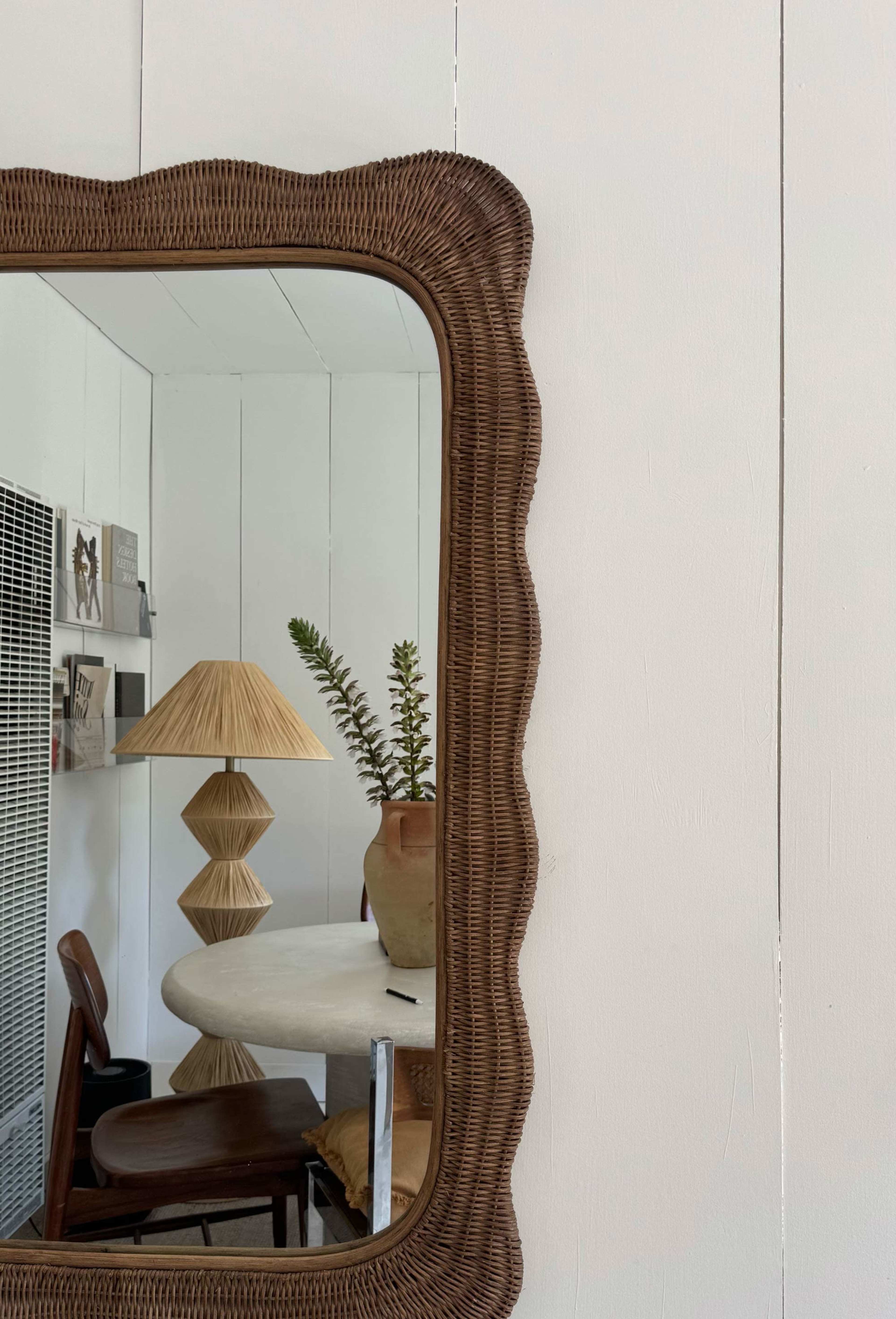 A decorative mirror with a scalloped frame reflects a lamp and a plant on a round table against a white wall.