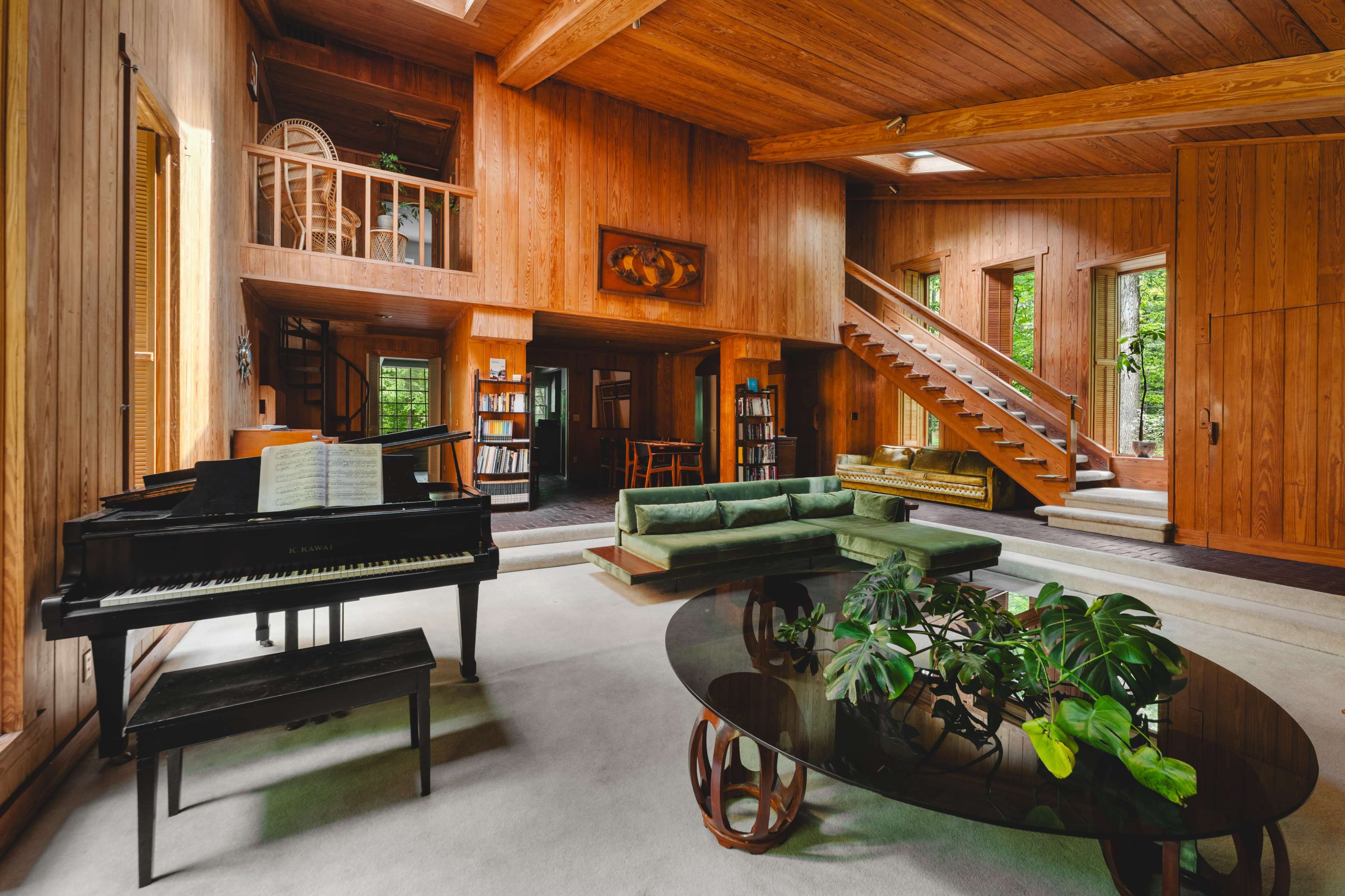 The image shows a spacious, wood-paneled living room with a piano, a green sectional sofa, a round glass coffee table, and stairs leading to an upper level.