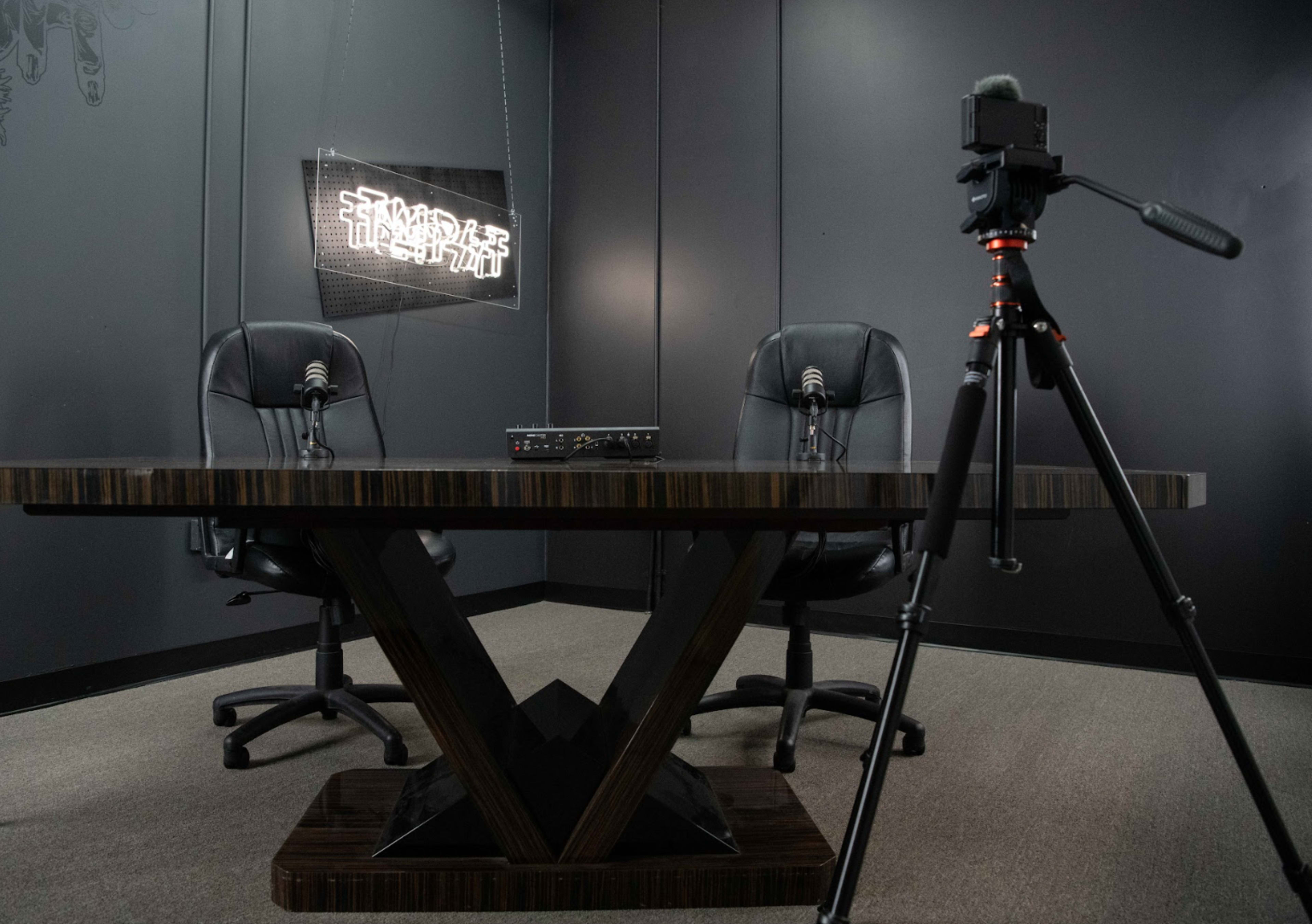 A recording studio setup with two black chairs facing a central table, a microphone on each chair, and a camera mounted on a tripod nearby.