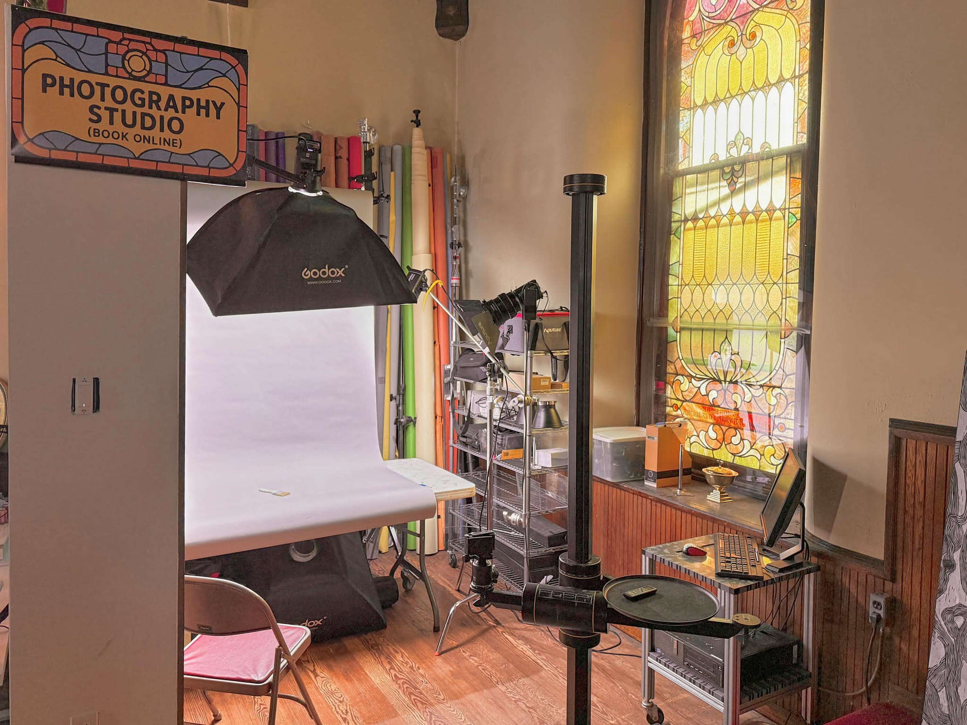 The image shows a photography studio with a backdrop setup, lighting equipment, and a colorful stained glass window.
