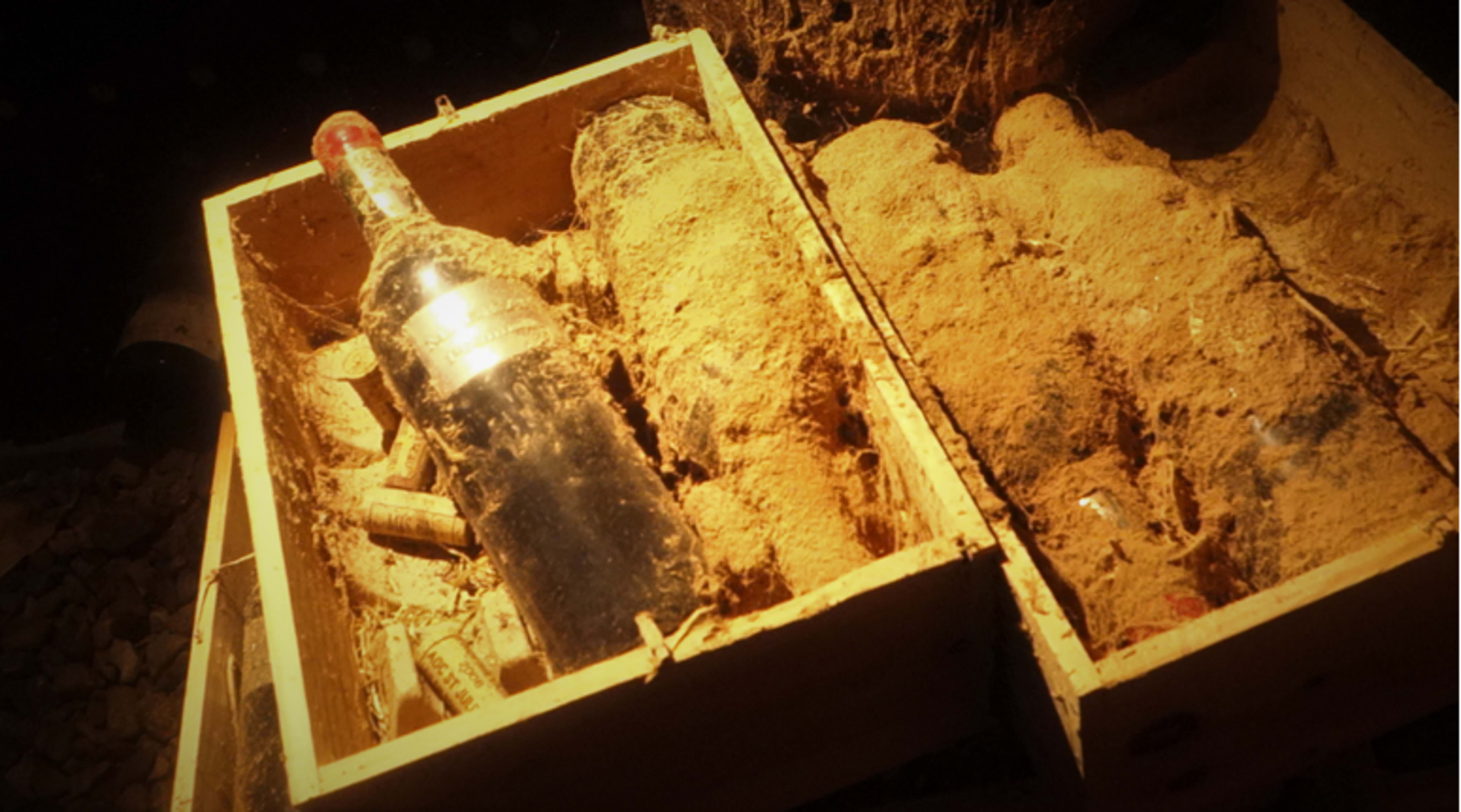 A wine bottle and several corked bottles are partially buried in dust inside a wooden crate.