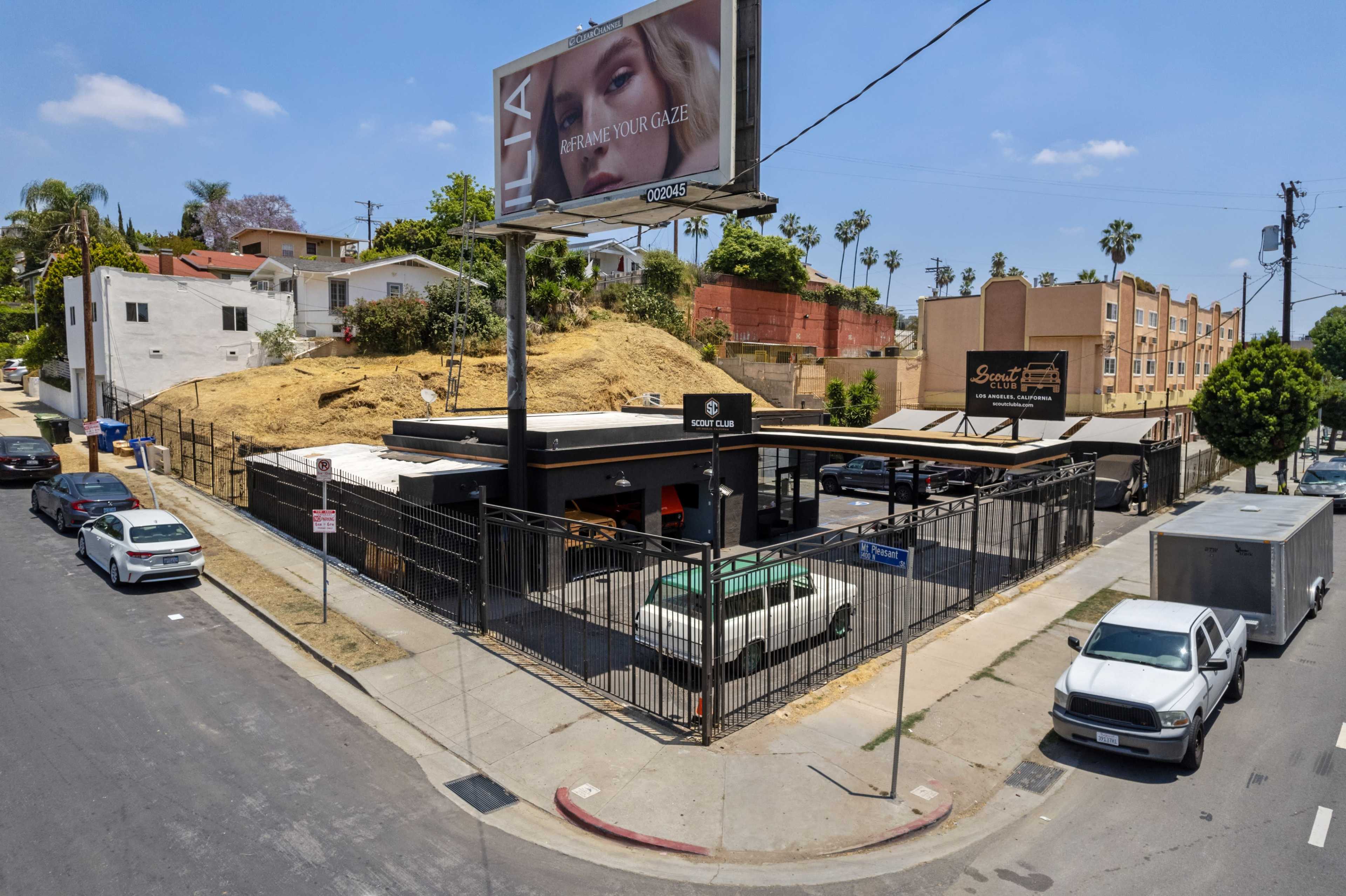 A fenced commercial building with a large billboard situated on a sloped lot, surrounded by vehicles parked on the street.
