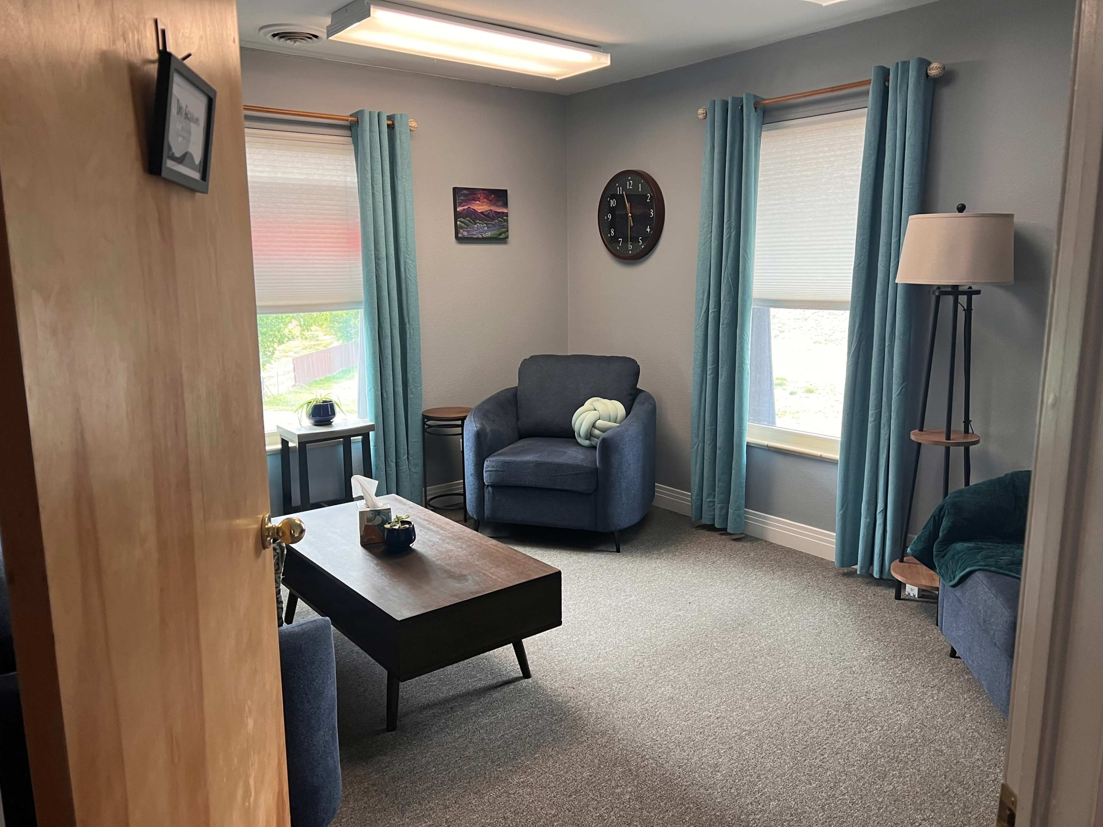 The image shows a cozy room with two windows, blue curtains, a clock on the wall, a blue armchair, and a coffee table in the center.