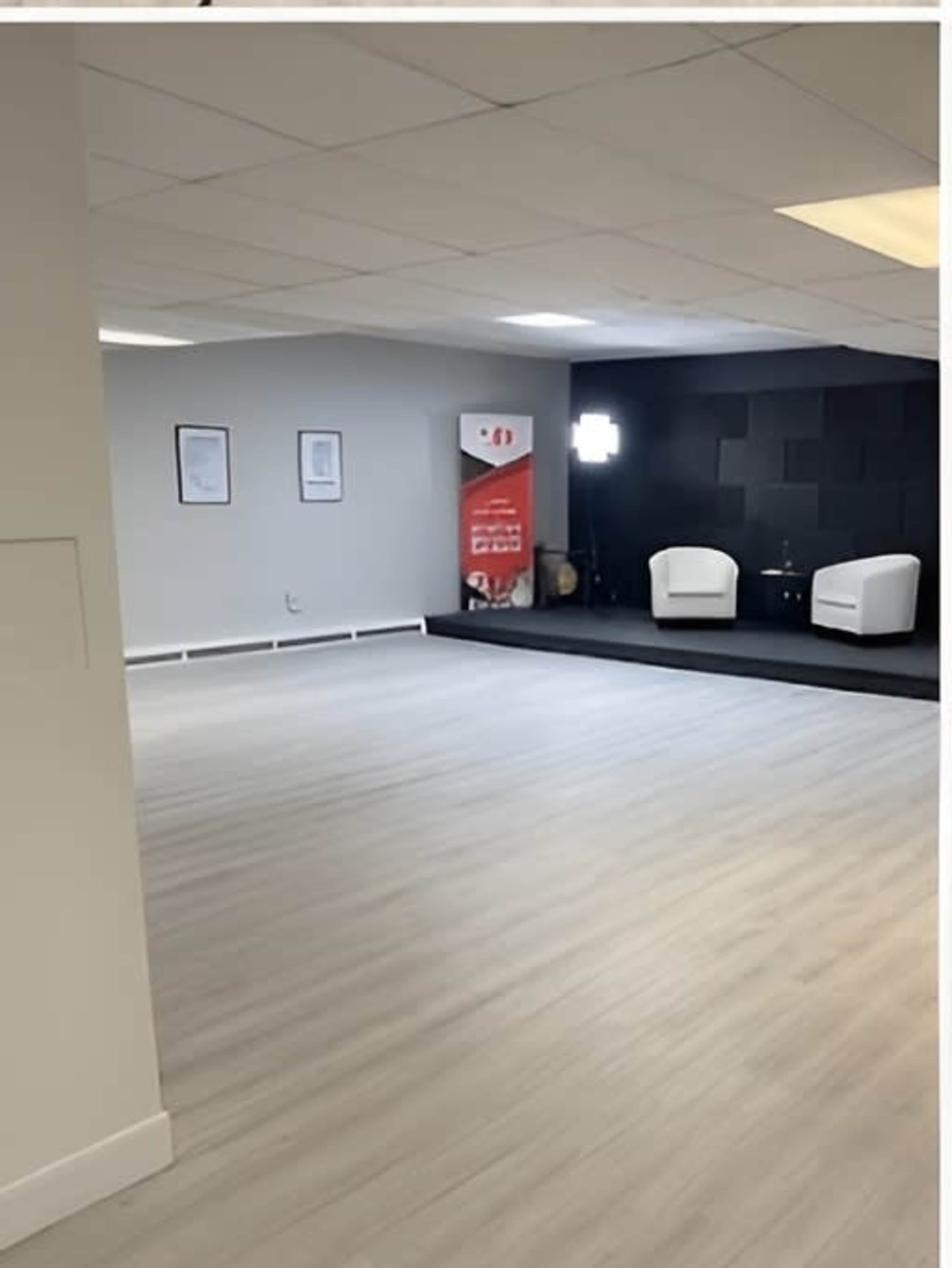 The image shows an empty room with light-colored flooring, featuring two white chairs on a black mat and a banner in the background.