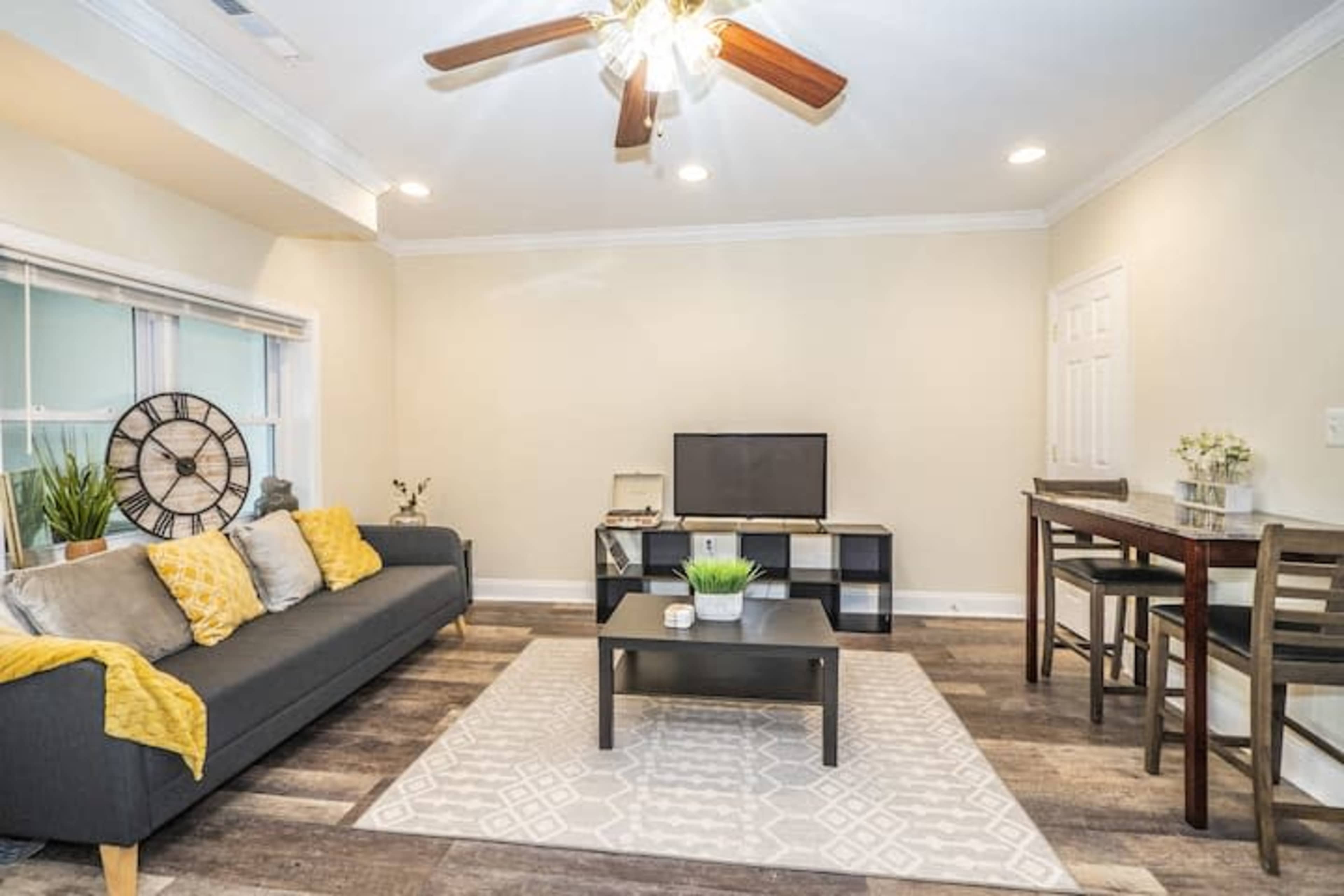 A living room features a dark sofa with yellow throw pillows, a coffee table, and a television on a stand against a light-colored wall.