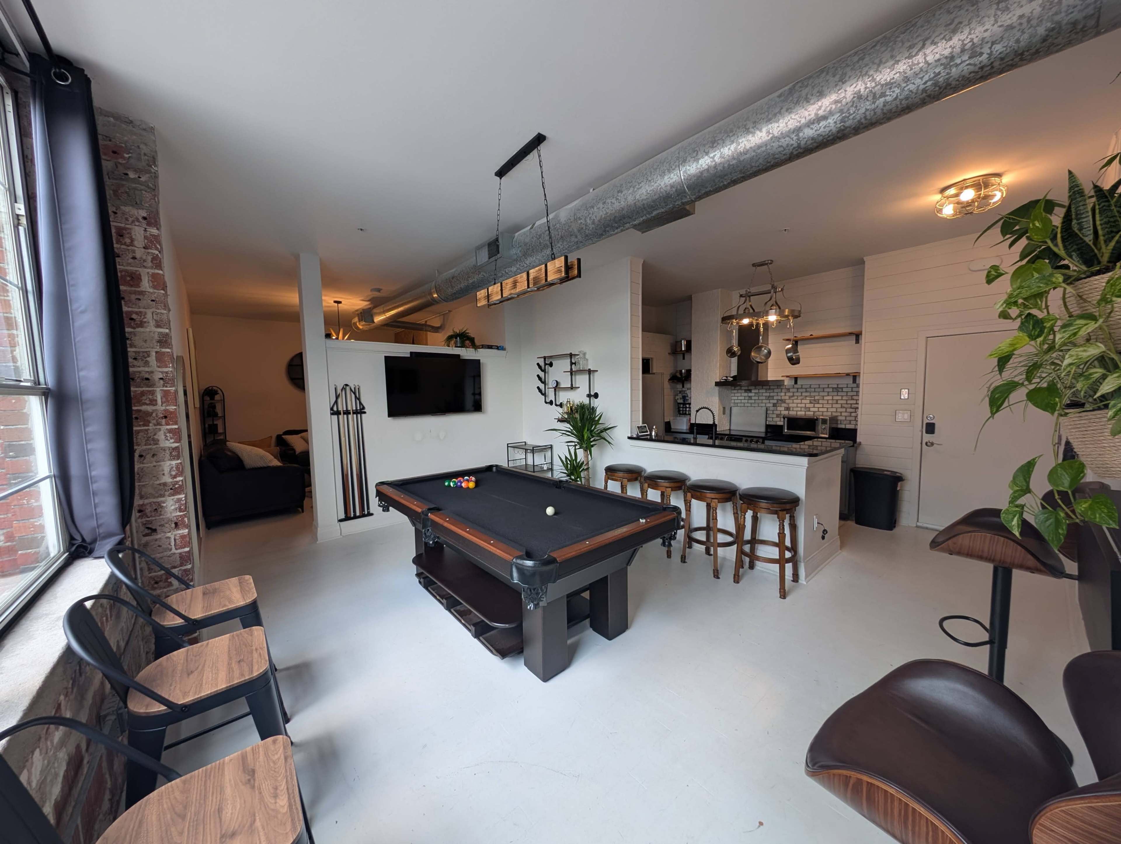 The image shows a spacious interior featuring a billiard table in the center, with seating areas, a kitchen counter, and large windows providing natural light.