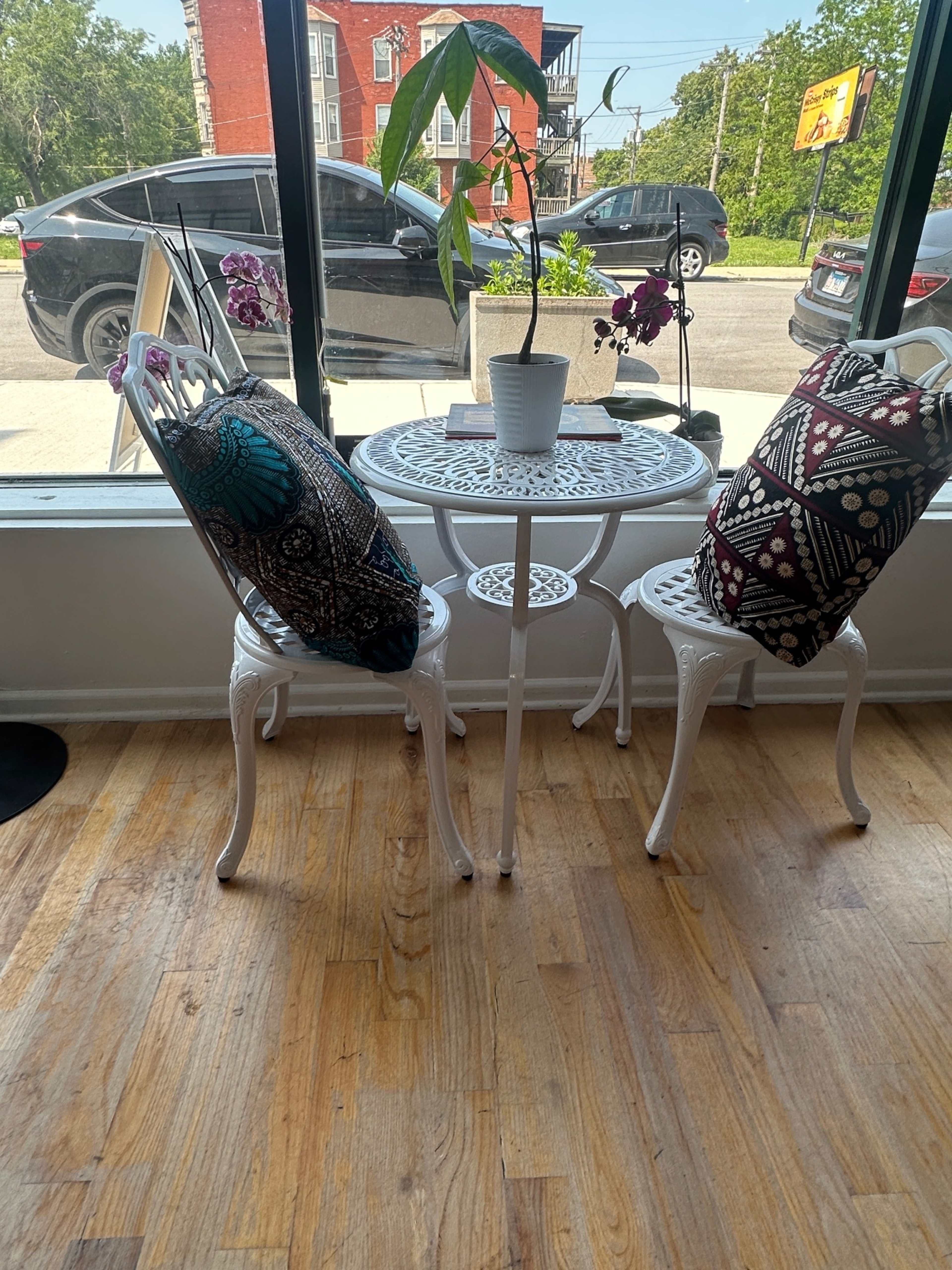 A small round table with two ornate white chairs, each adorned with decorative pillows, is positioned near a window with a potted plant and orchids on top.