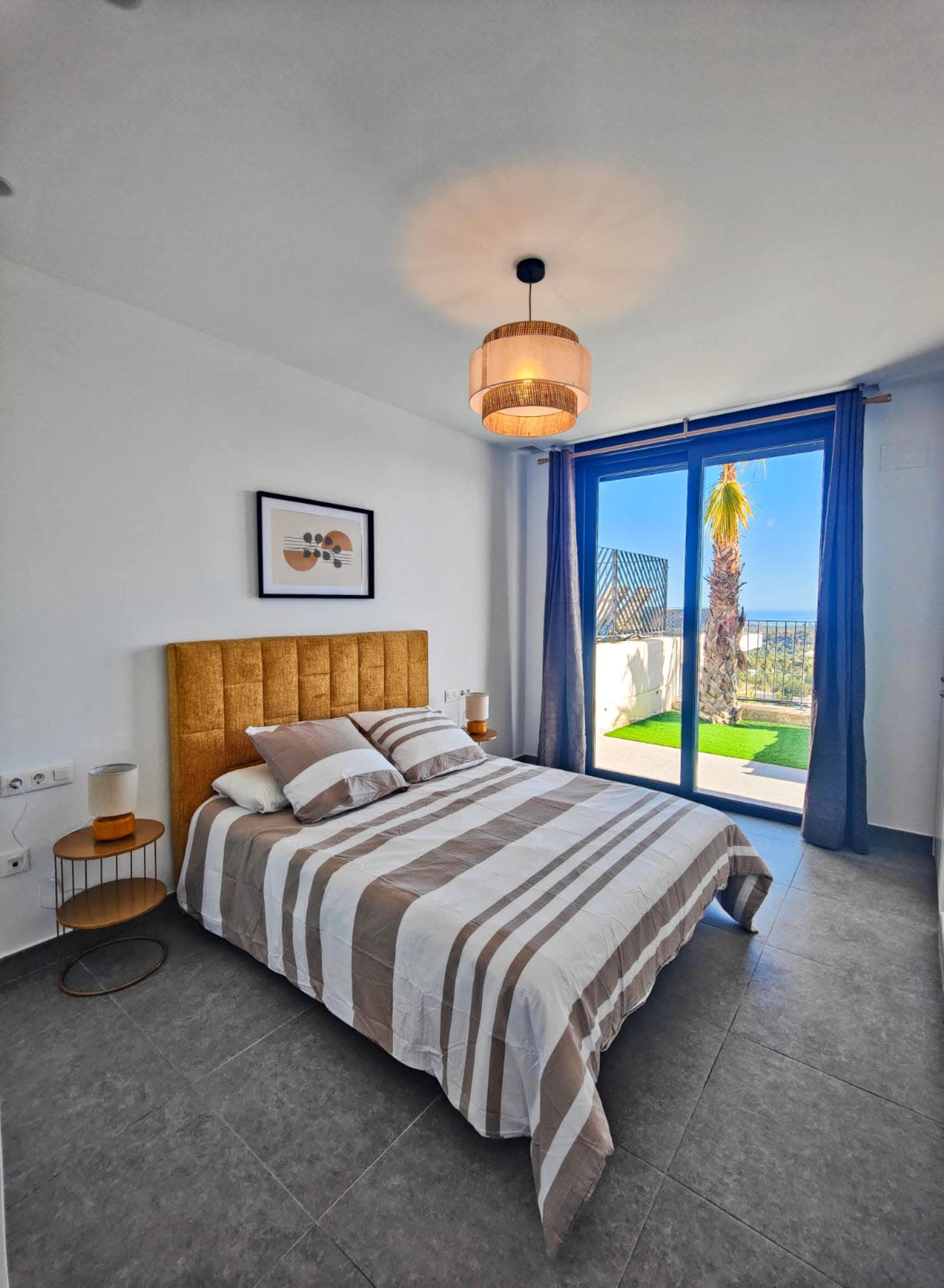 The image shows a modern bedroom with a striped bedspread, two bedside tables, and large glass doors leading to a terrace with a view of palm trees.