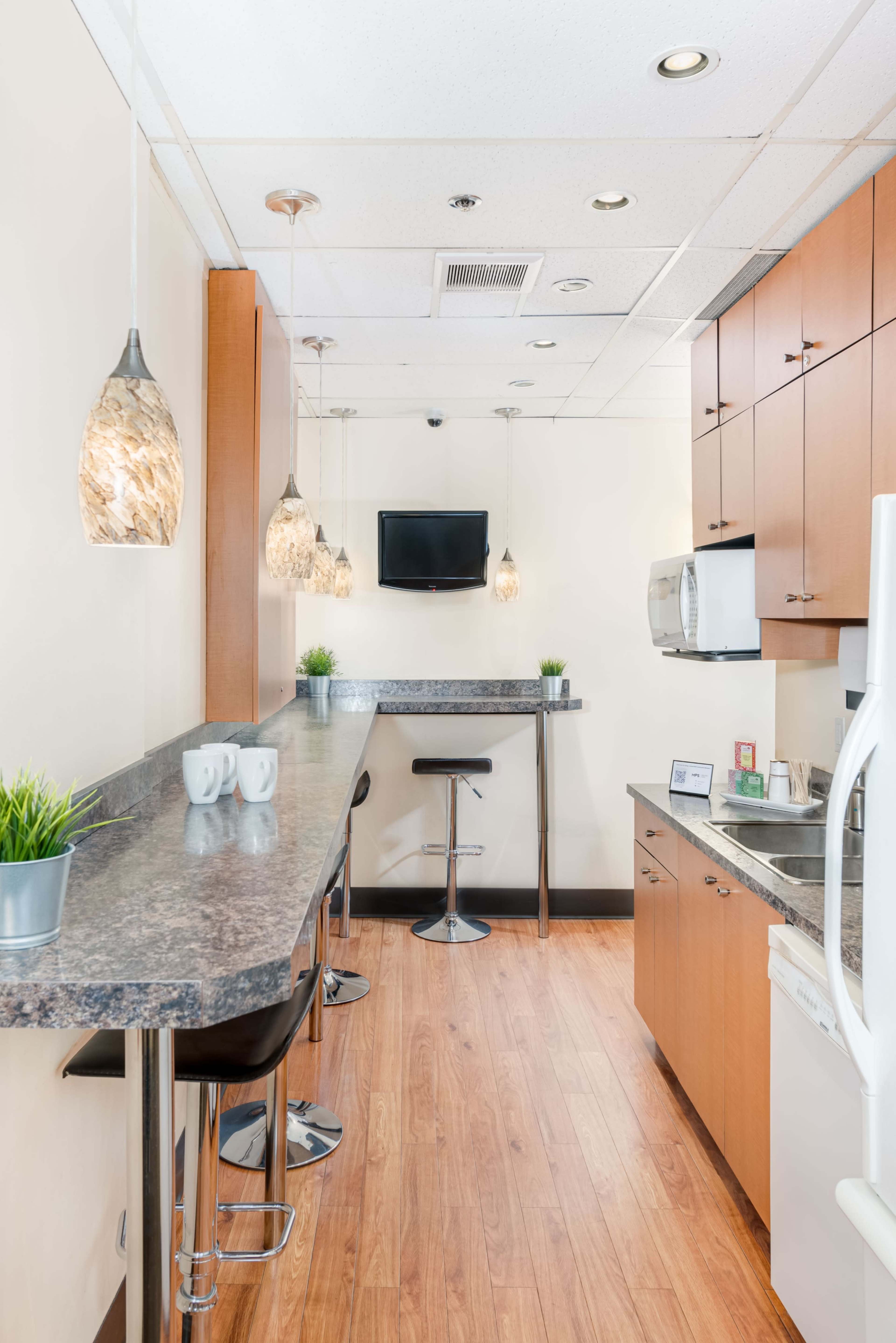 The image depicts a modern kitchen area with a countertop bar, stools, and pendant lights, alongside appliances and cabinets in a bright, spacious environment.