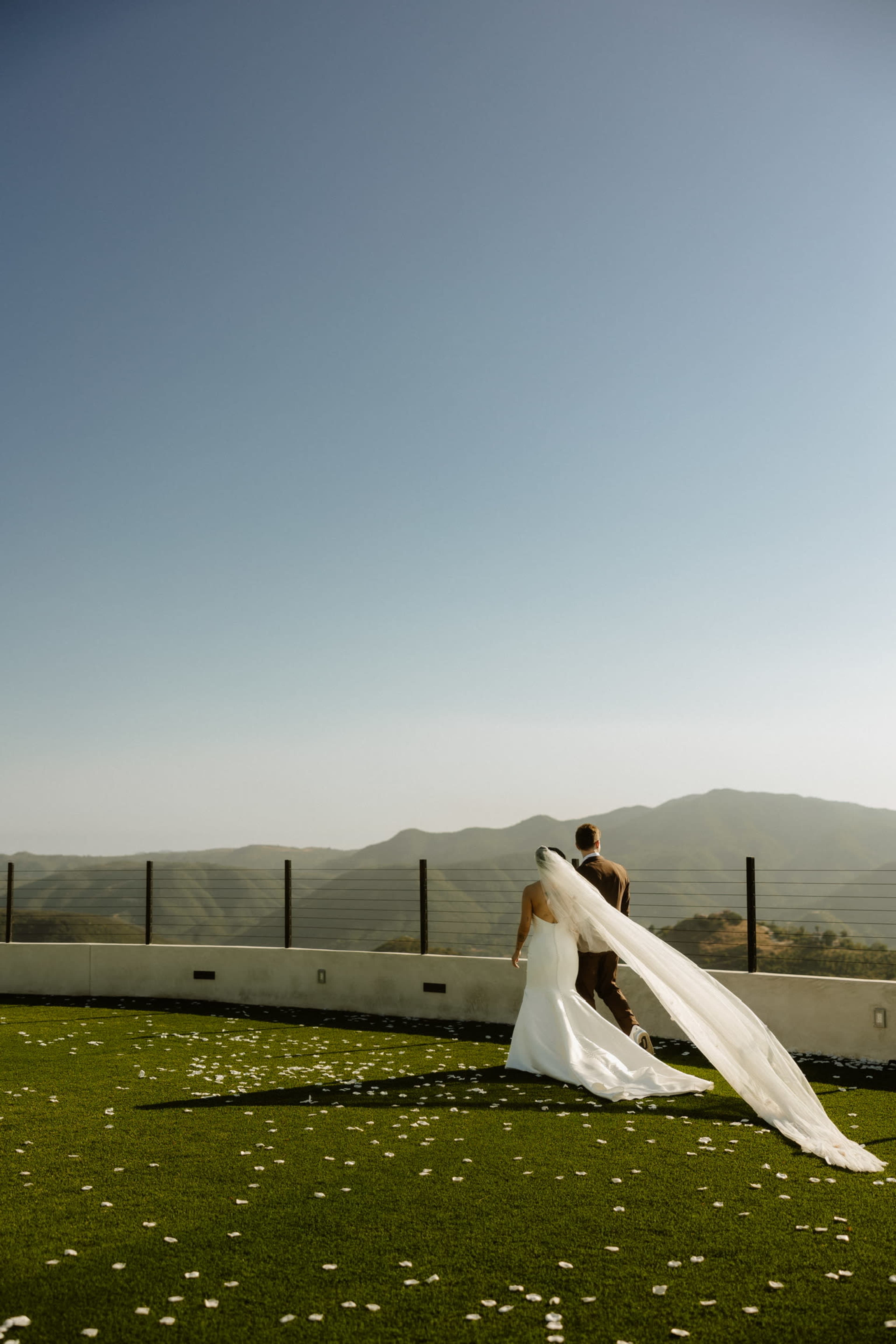 A couple walks hand-in-hand on a green lawn overlooking mountains under a clear blue sky.