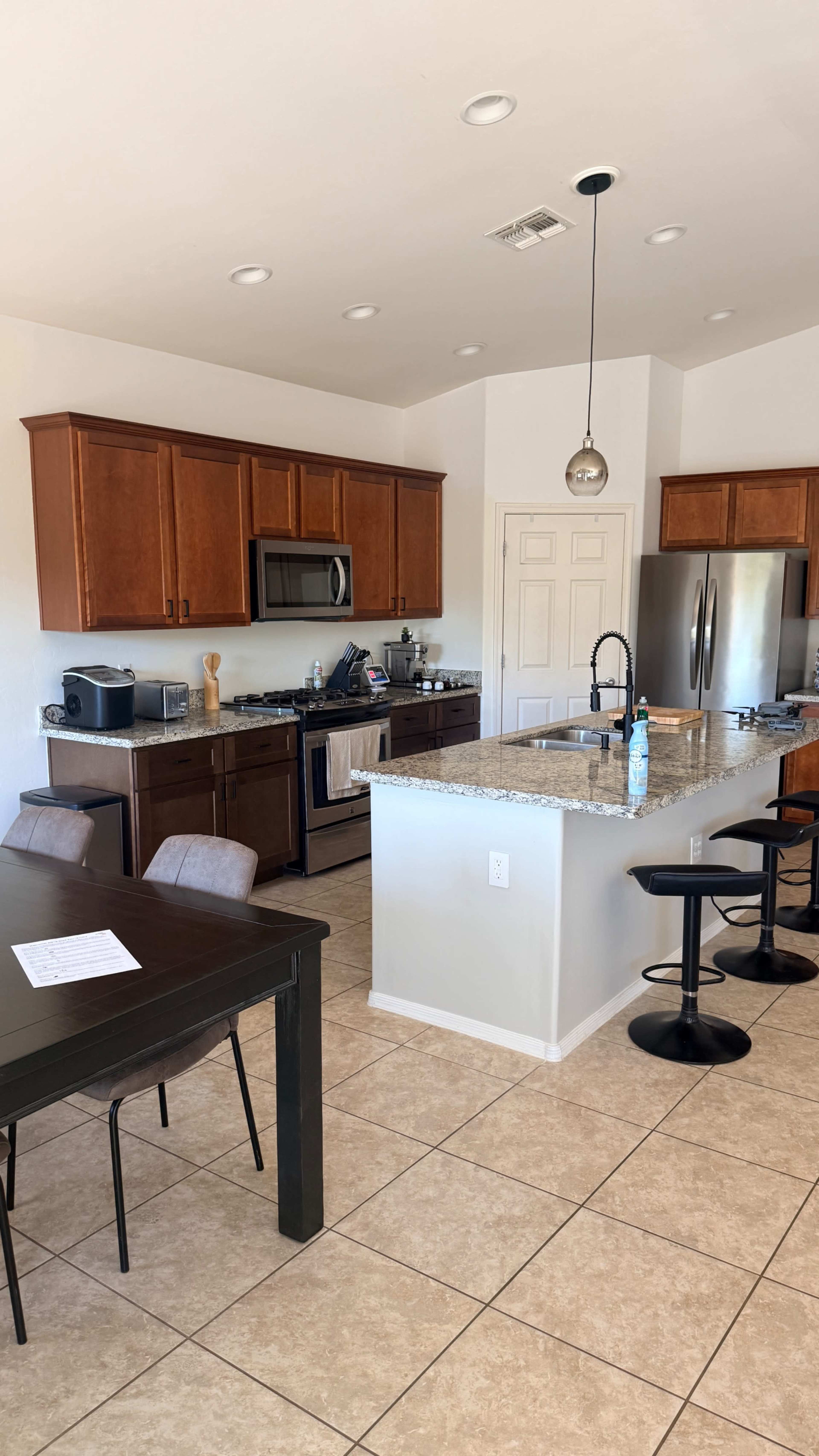 The image shows a modern kitchen with wooden cabinets, stainless steel appliances, and a central island with bar stools, along with a dining table nearby.