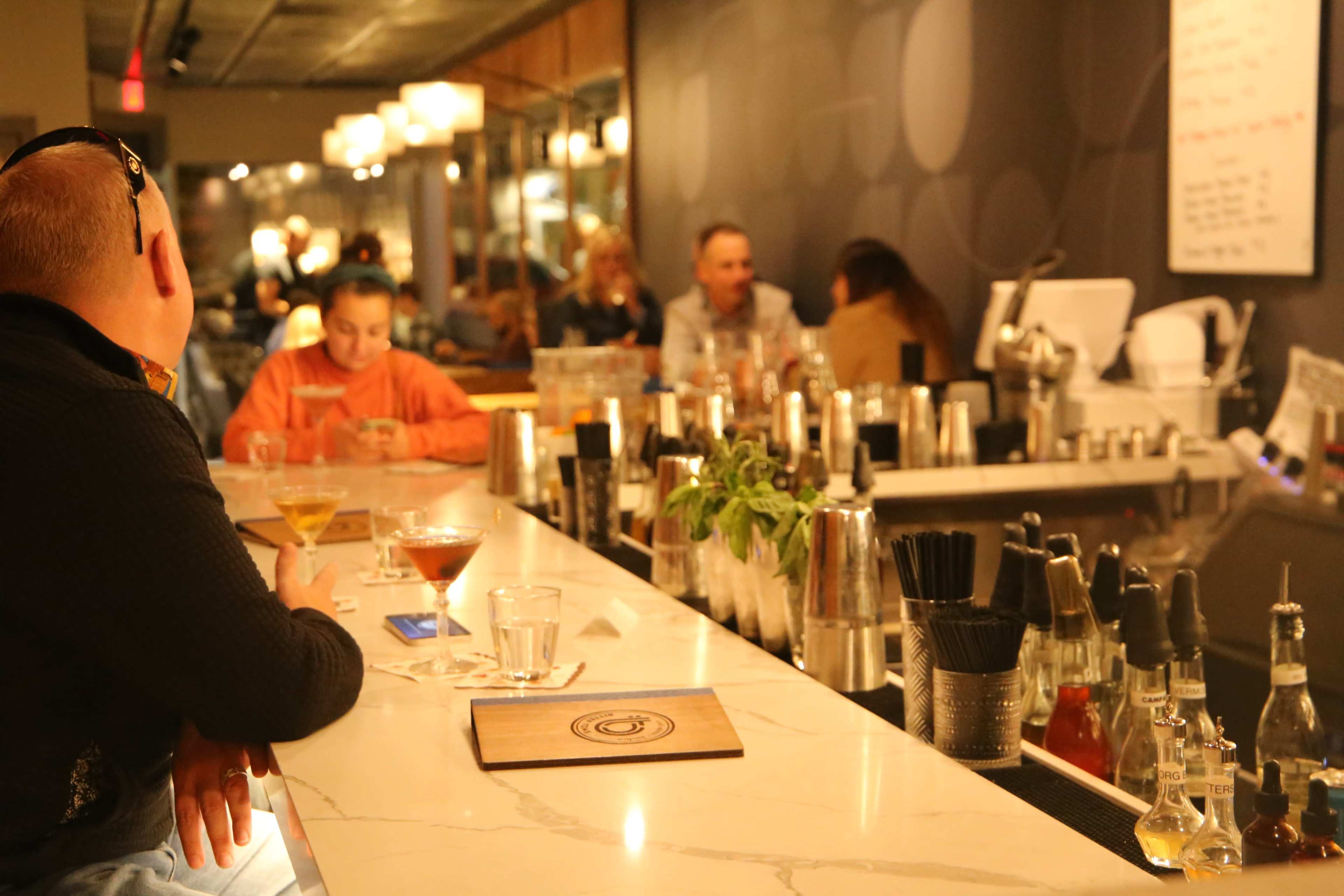 A bar scene shows patrons seated at a marble countertop while others are engaged in conversation in the background.