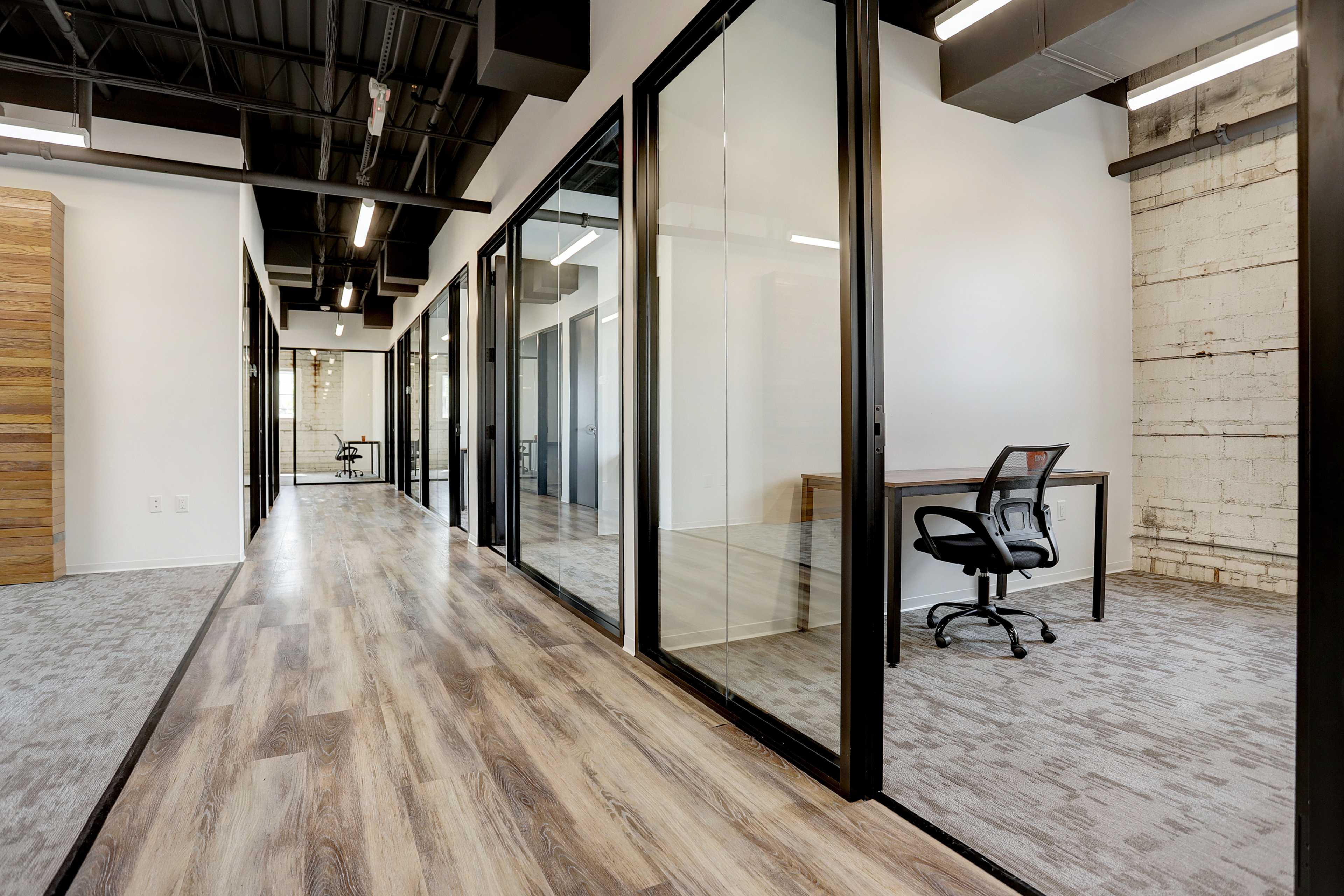 A modern office interior featuring glass-walled meeting rooms along a corridor with wooden and carpeted flooring.