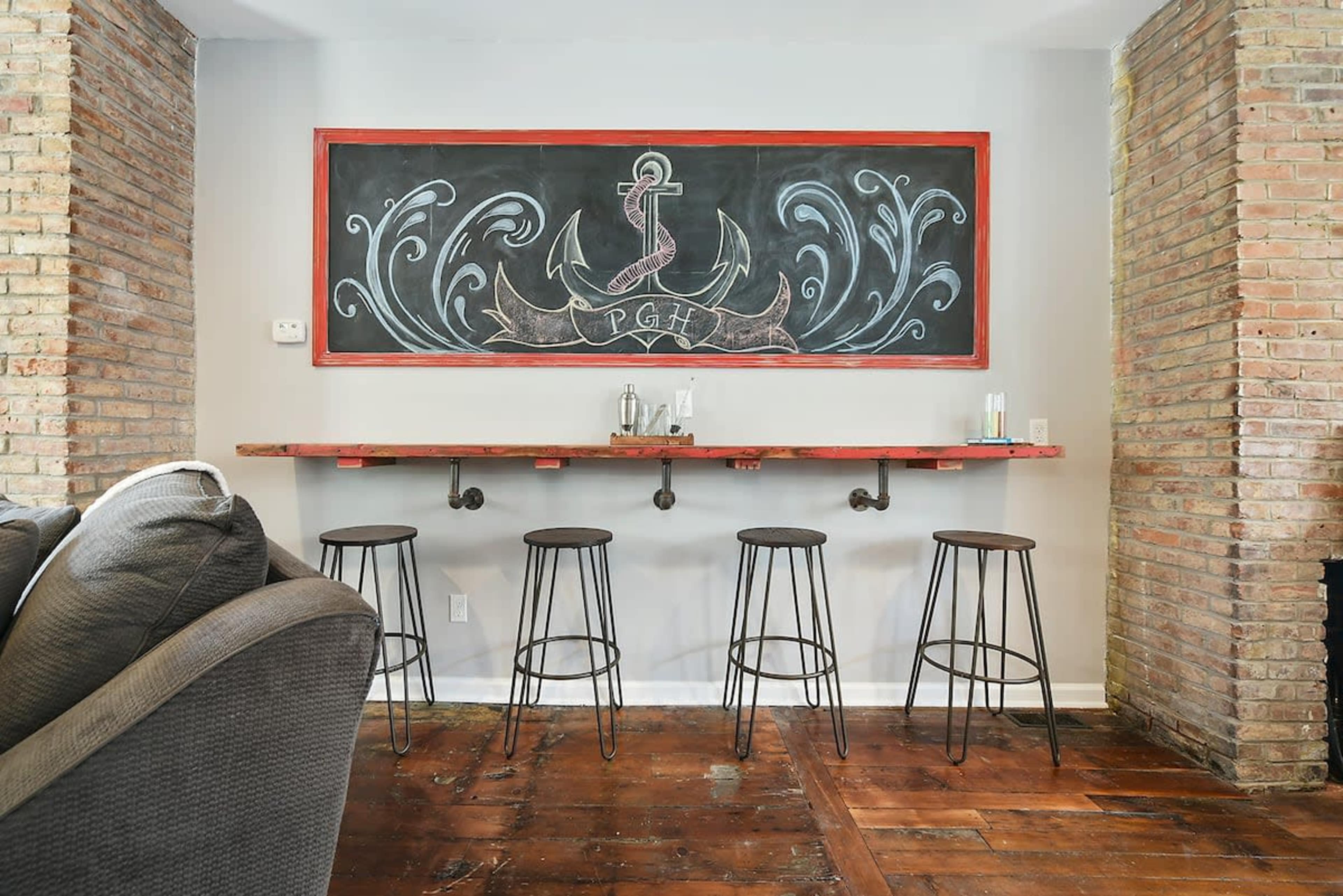 A chalkboard with decorative designs and the letters "PGH" is mounted above a rustic wooden bar with four metal stools.