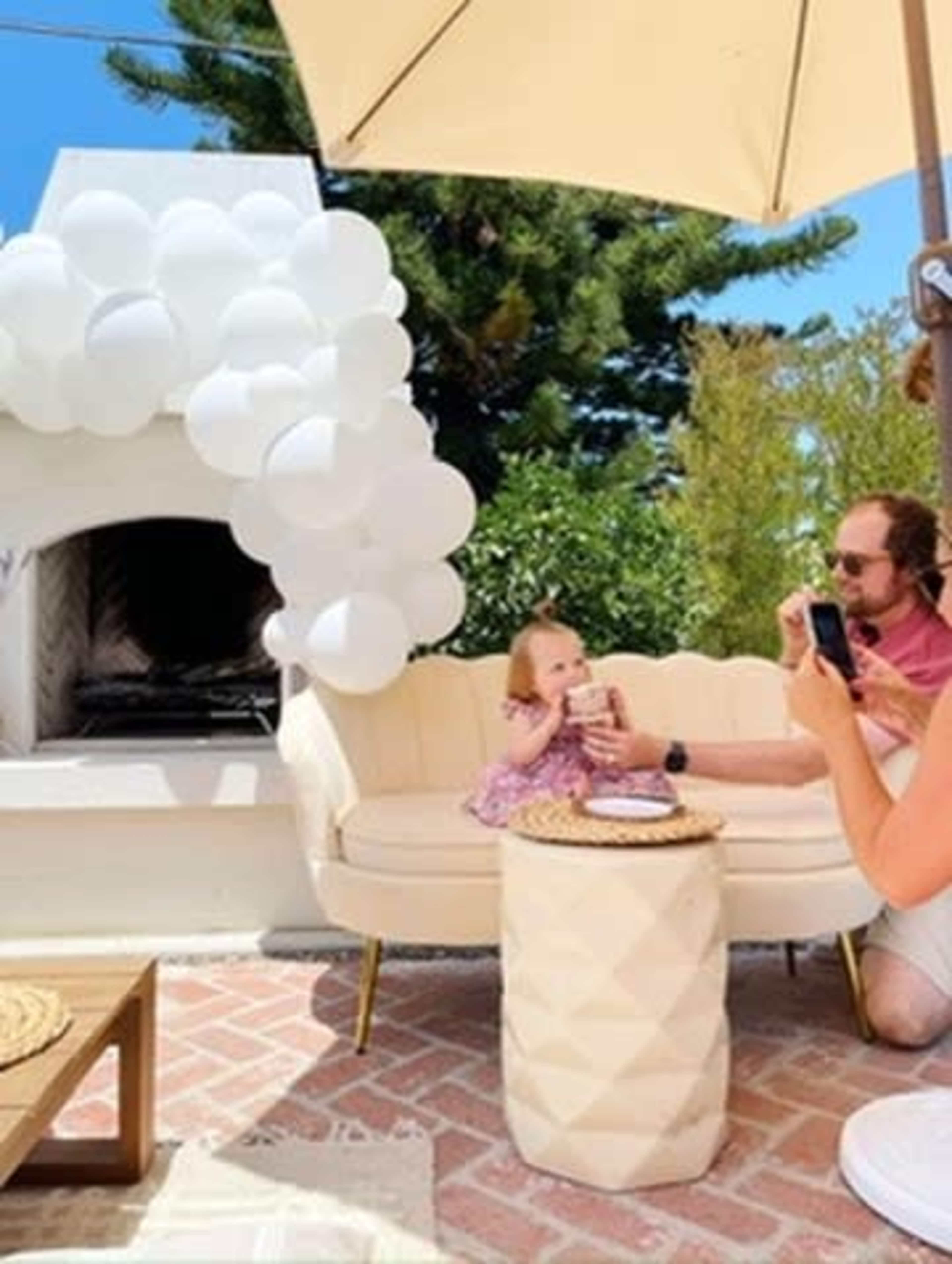 A child sits on a couch in a backyard setting while an adult takes a photo, with a decorative balloon arrangement and a fireplace in the background.