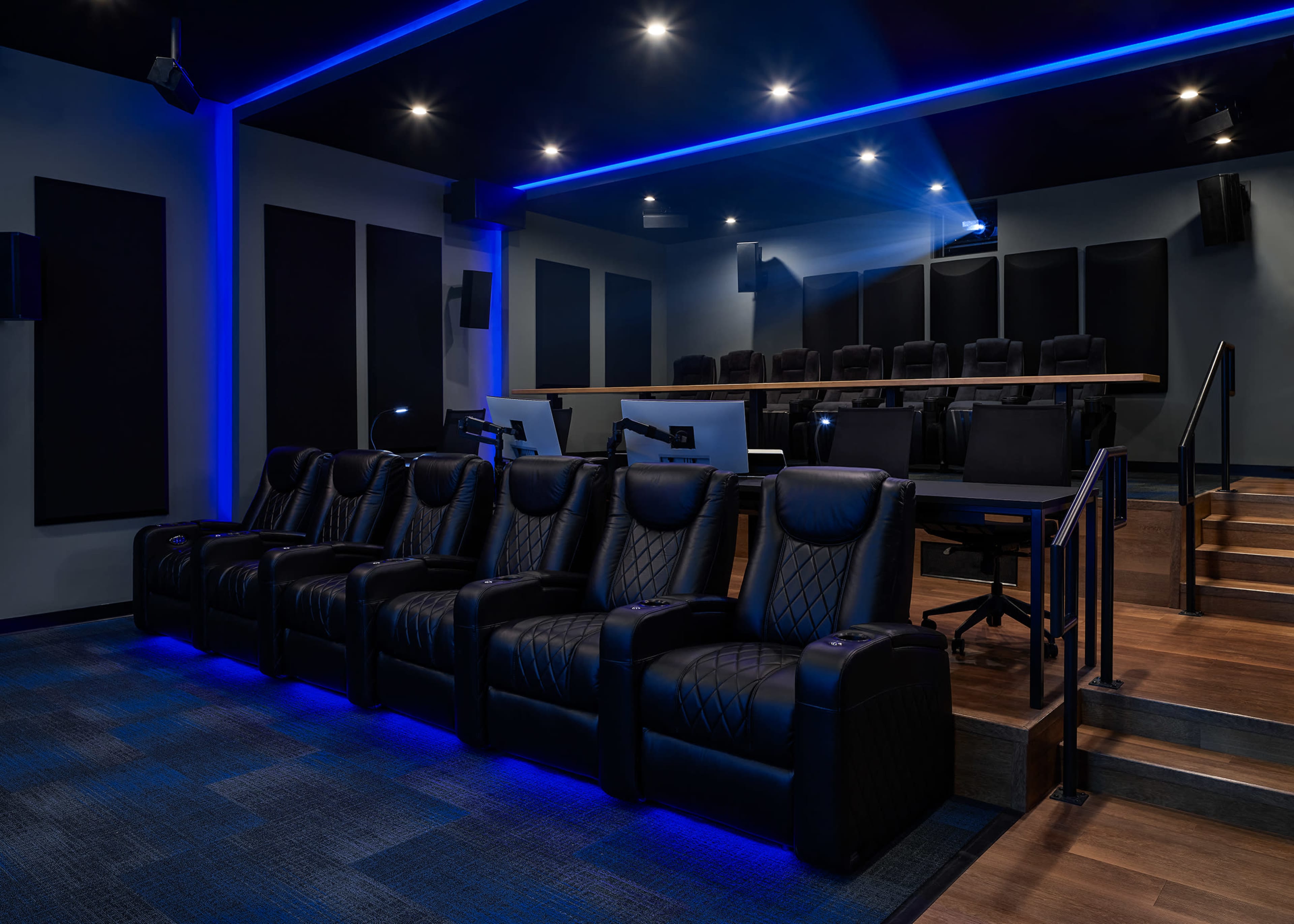 A modern home theater with black recliner seats arranged in front of a tiered seating area and ambient blue lighting.