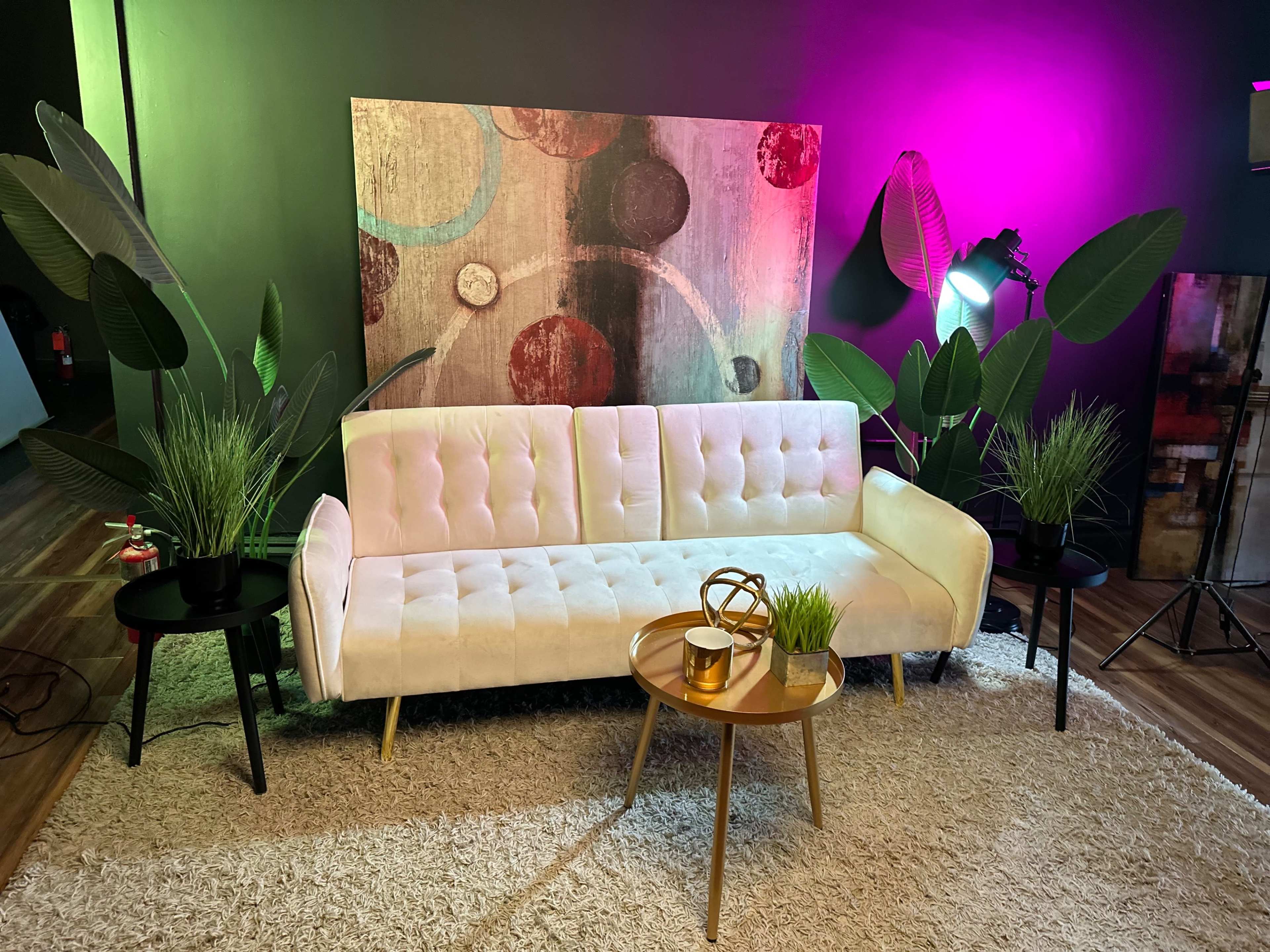 The scene features a white tufted sofa arranged with two black side tables and a round coffee table, surrounded by decorative plants and a colorful abstract painting on the wall.