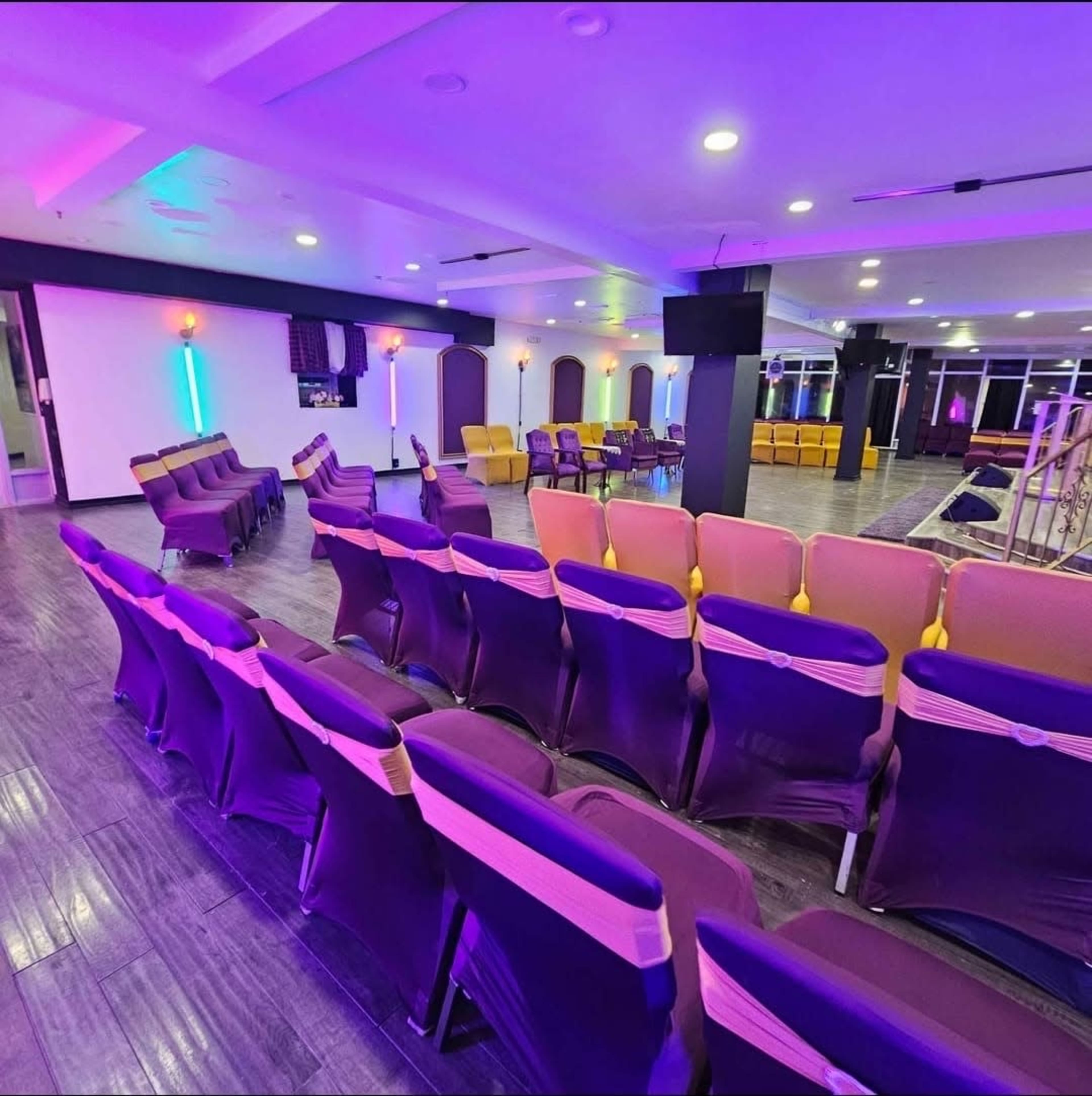 The image shows a spacious indoor venue with rows of purple and yellow seating arranged in a semi-circular layout, illuminated by colorful lights.