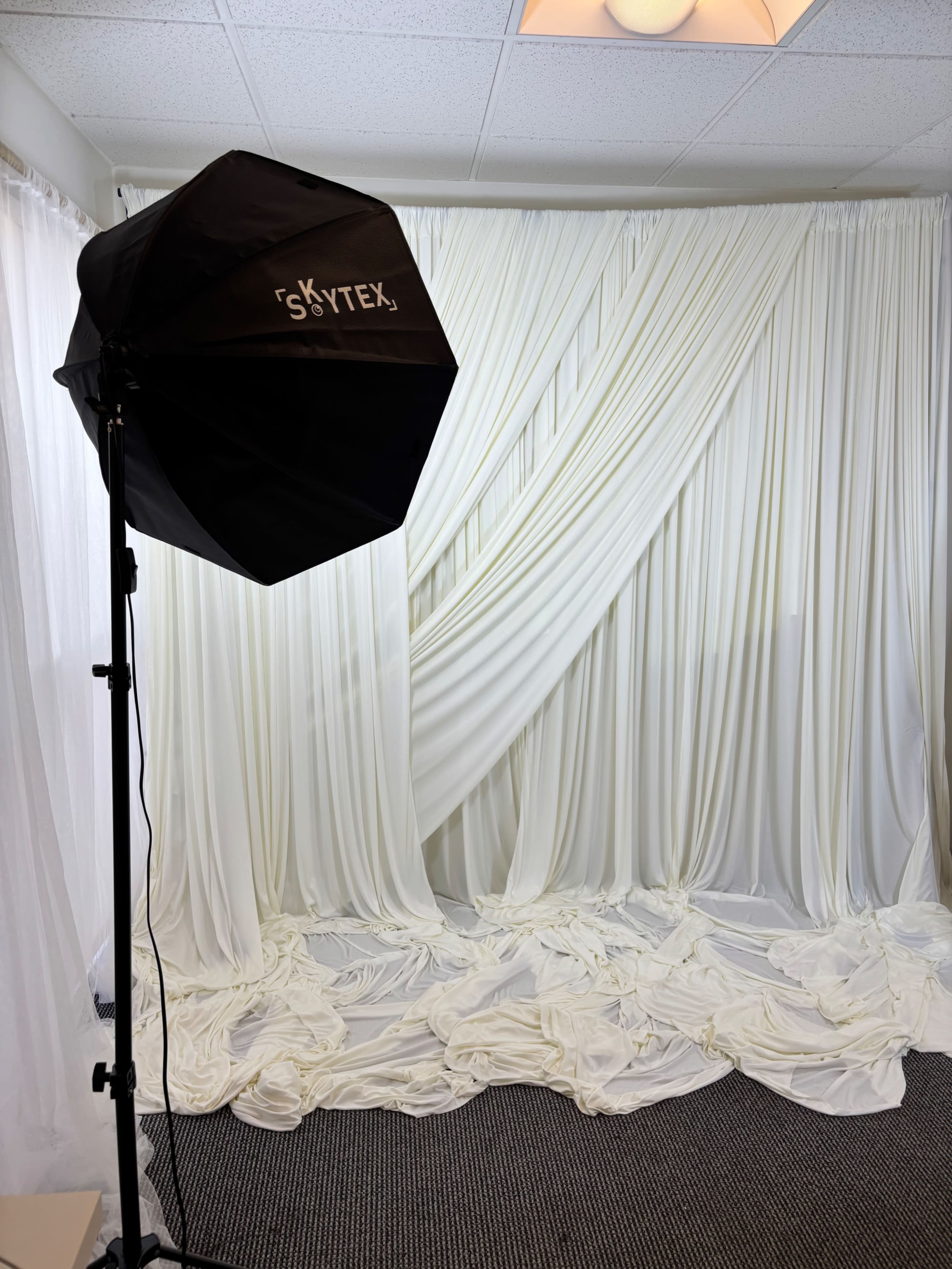 The image shows a photo studio setup with a softbox light on a stand beside a backdrop of flowing white drapery on the floor.