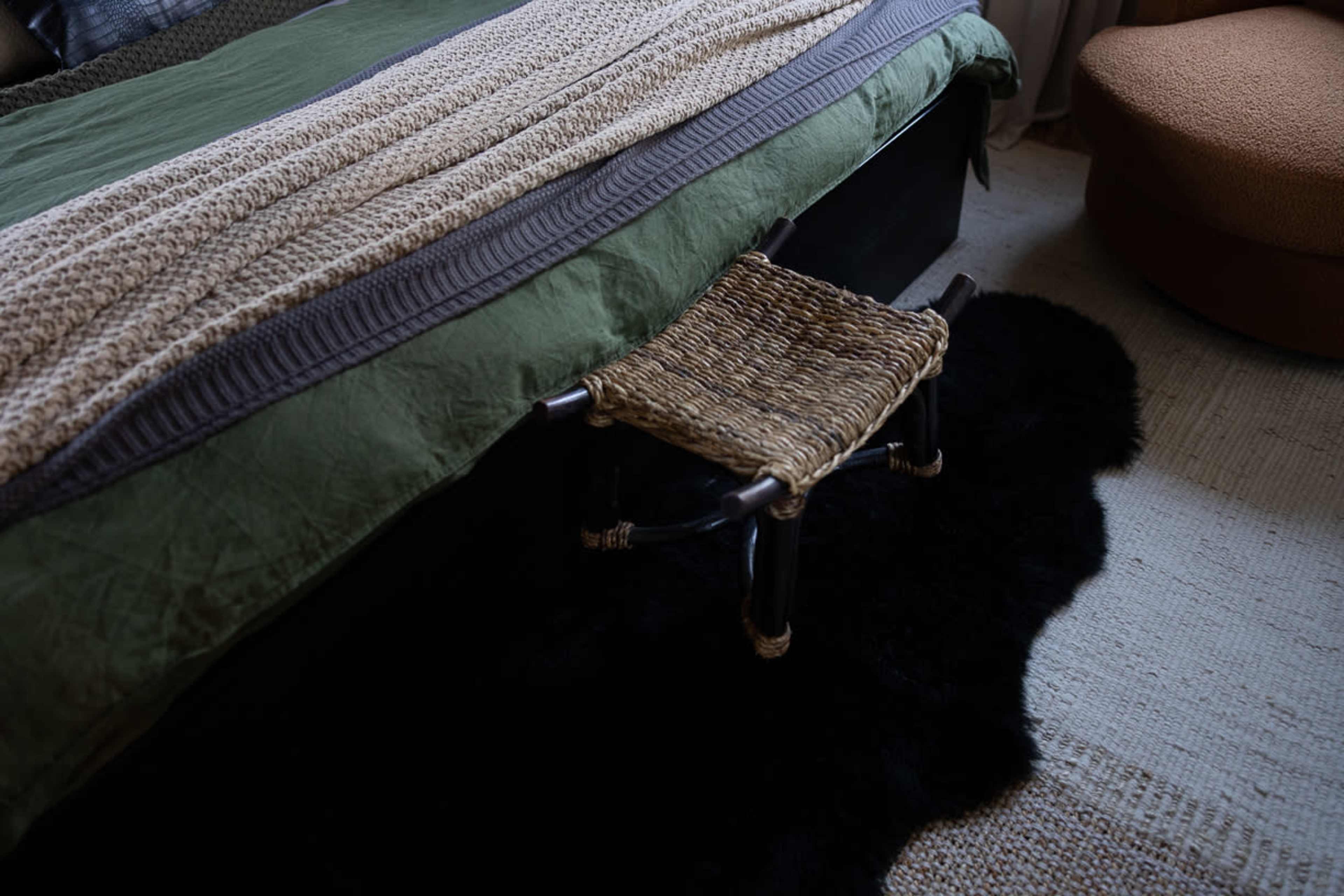 A woven footstool rests beside a bed covered with a green quilt and a textured blanket, on a dark rug.