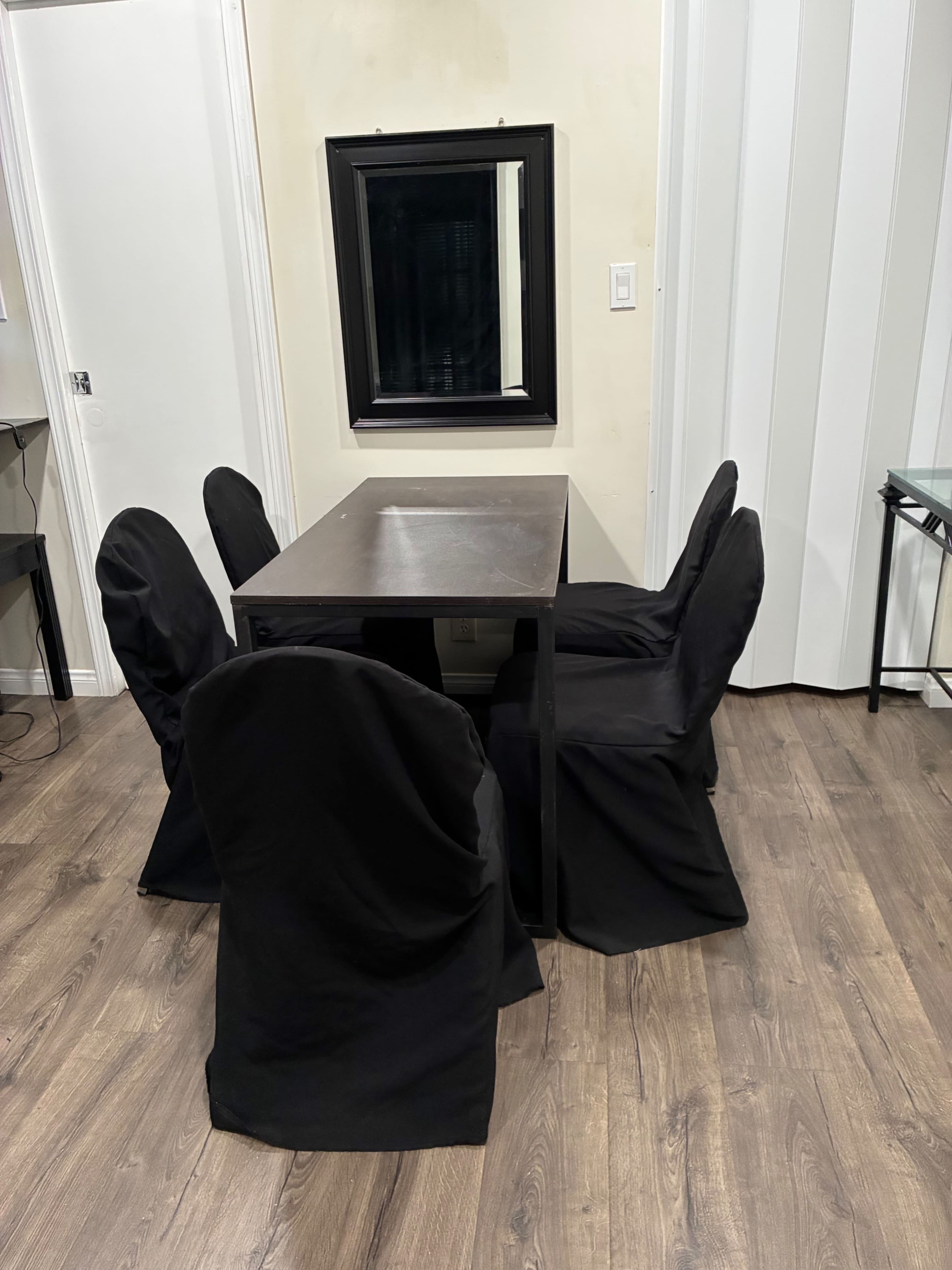 A rectangular dining table surrounded by four chairs, all covered with black fabric, is positioned against a wall with a mirror above it.