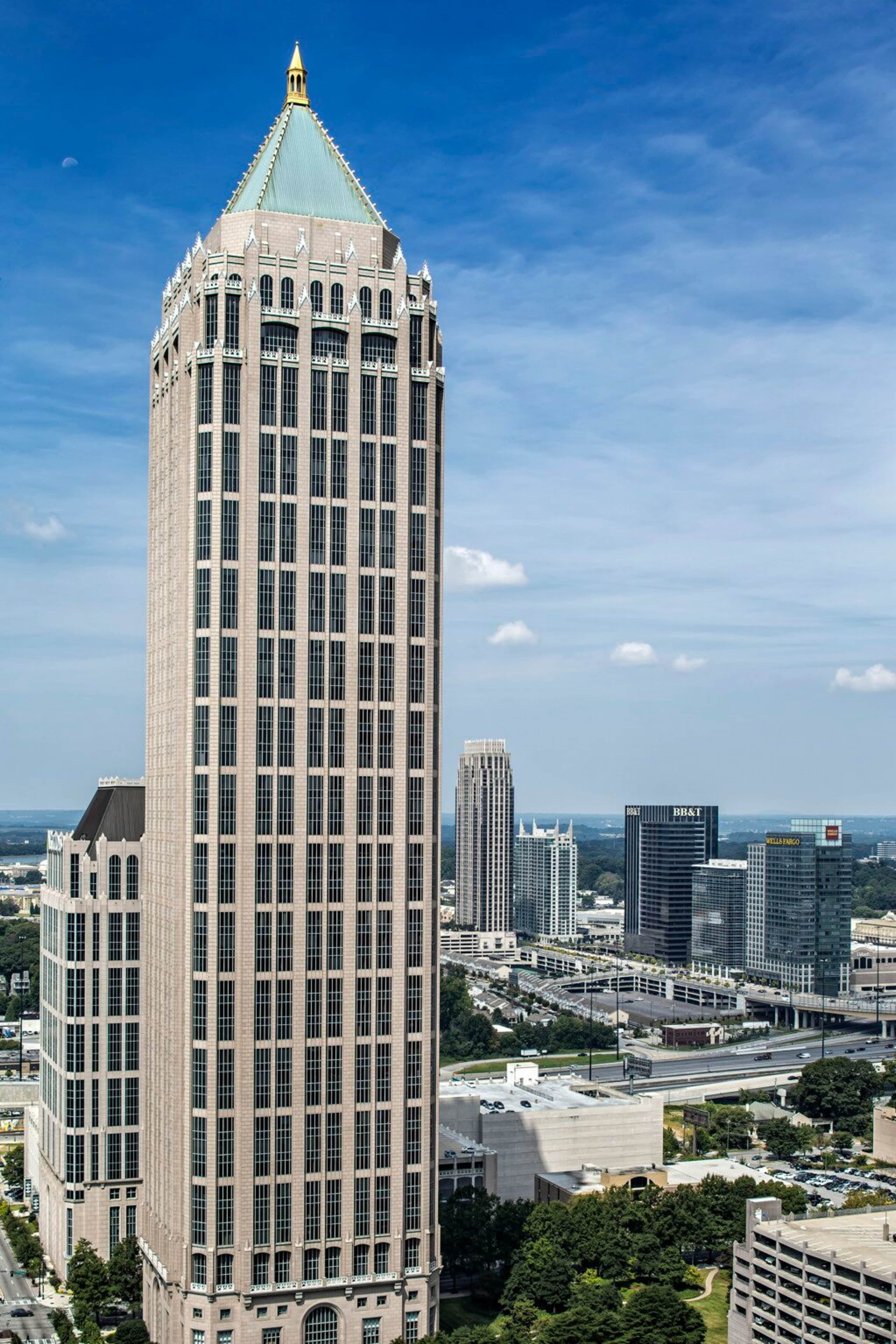 A tall skyscraper with a pointed roof stands prominently against a blue sky, surrounded by other buildings in an urban landscape.