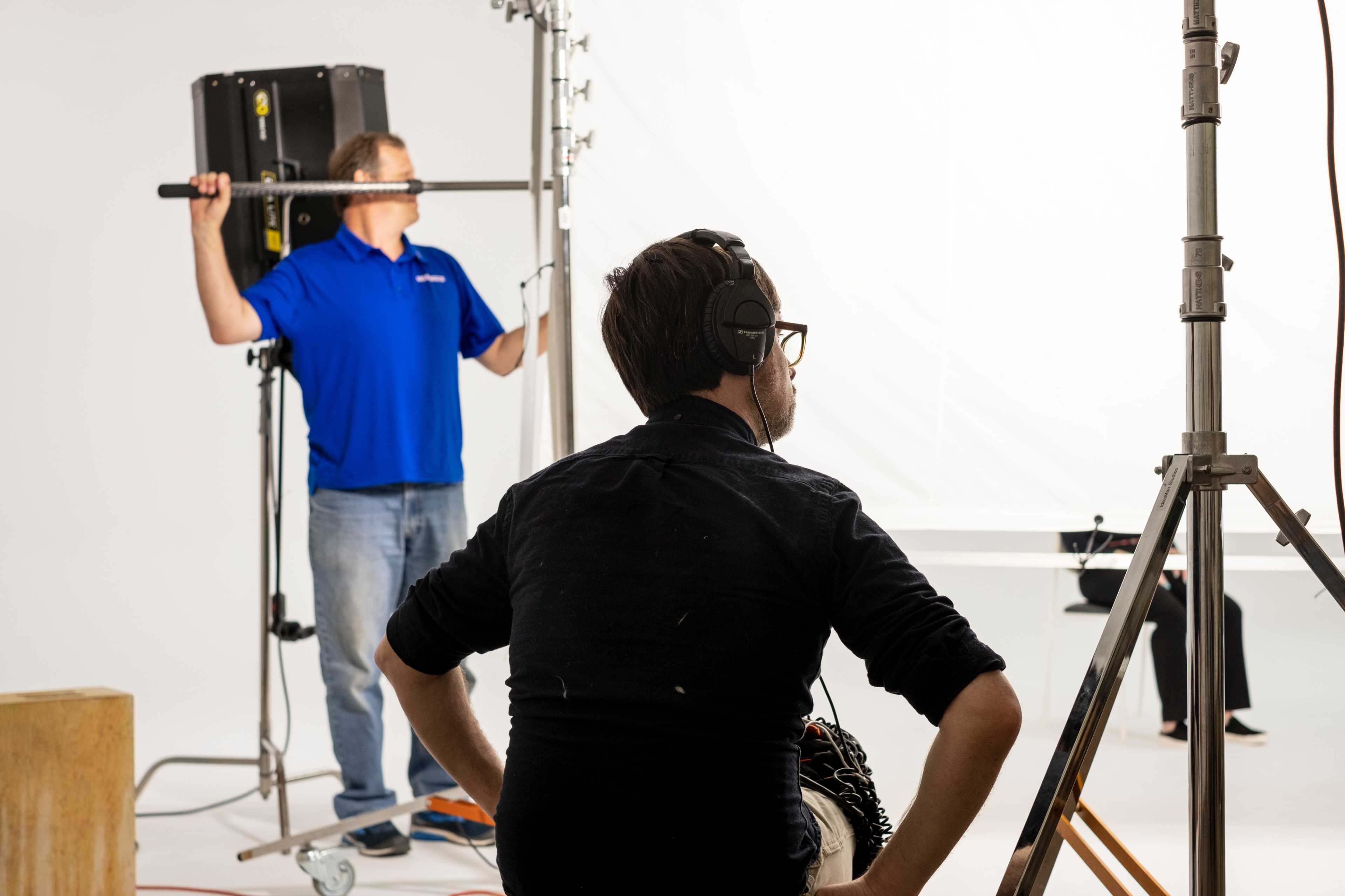 A person sits in front of a camera setup while another person adjusts a lighting rig in a studio.