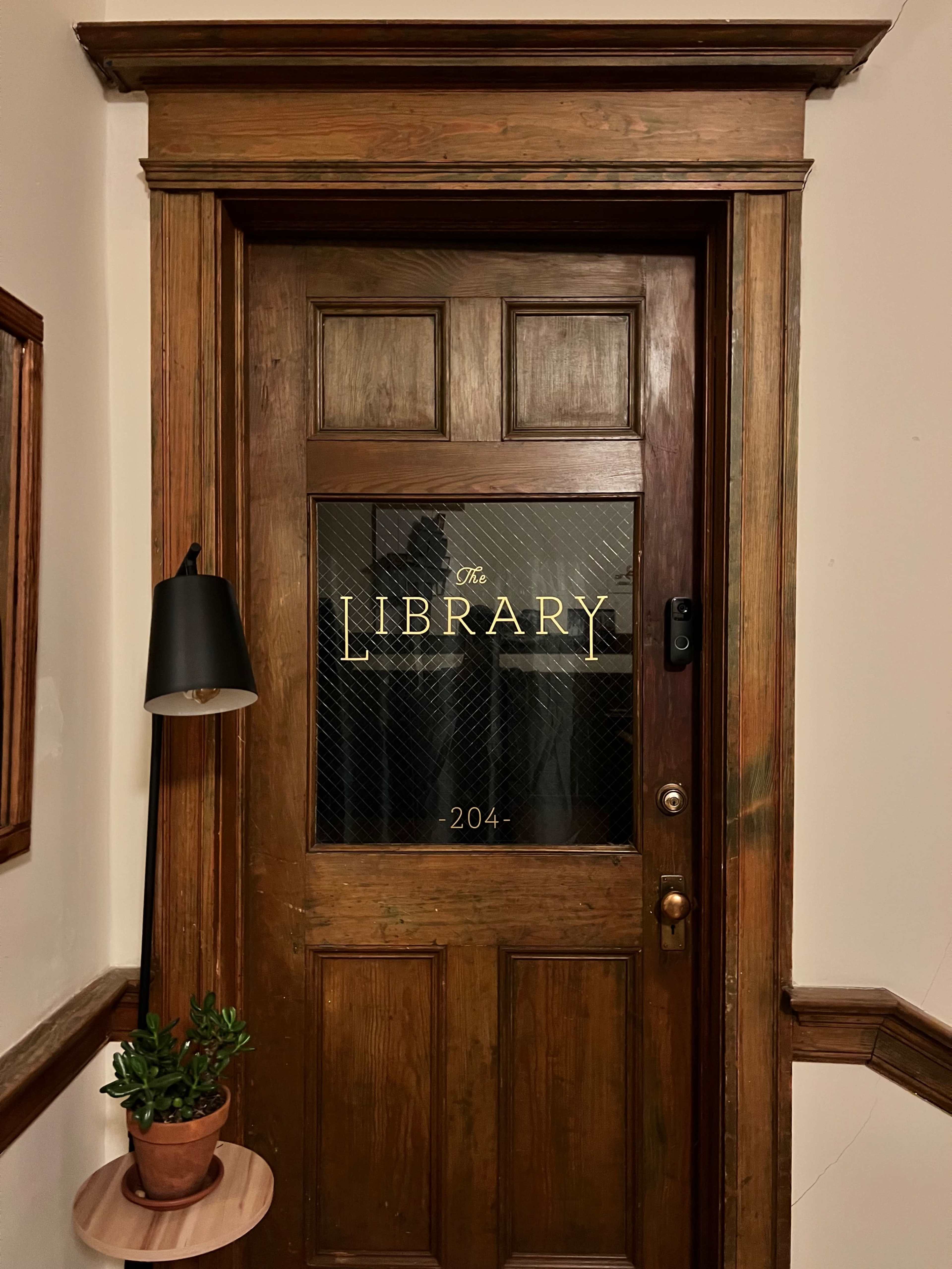 The photo shows a wooden door with a glass panel labeled "The Library" and the room number "204," complemented by a wall-mounted lamp and a potted plant on a small side table.