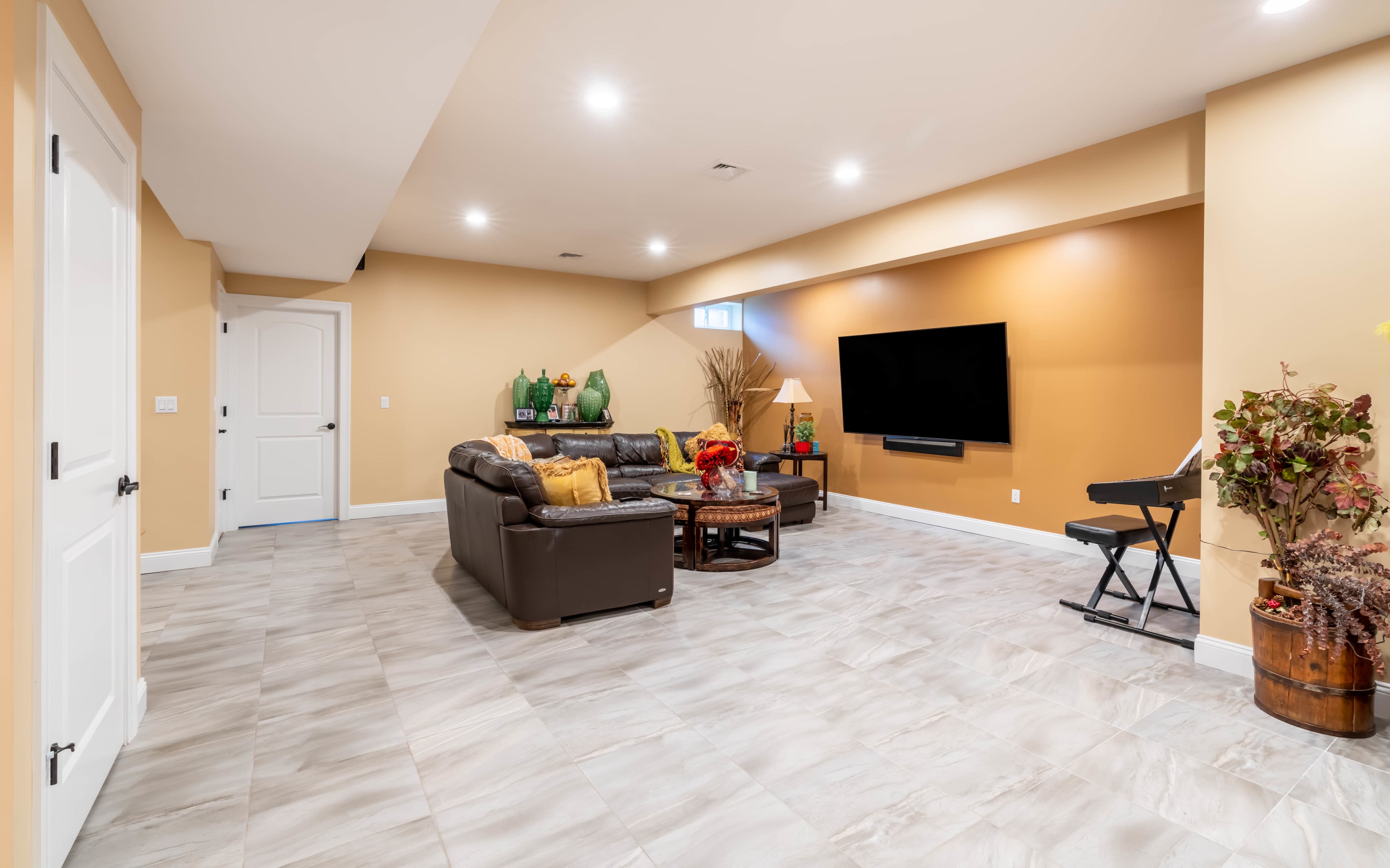 A spacious, well-lit basement features a seating area with a dark leather couch, a large flat-screen TV on the wall, and a small keyboard on a stand.