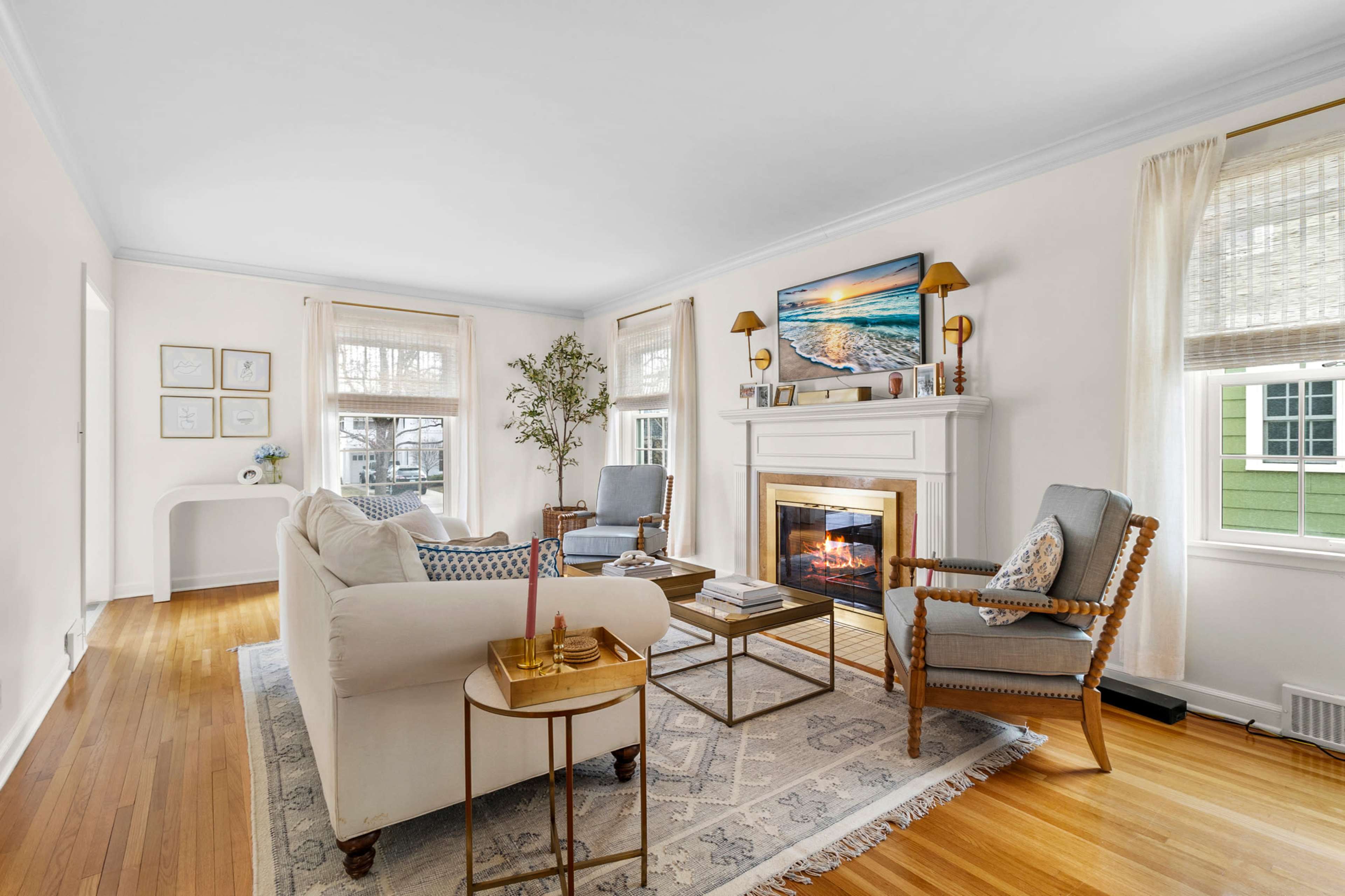The bright living room features a cozy fireplace, two armchairs, a sofa, and windows allowing natural light to illuminate the wood flooring.