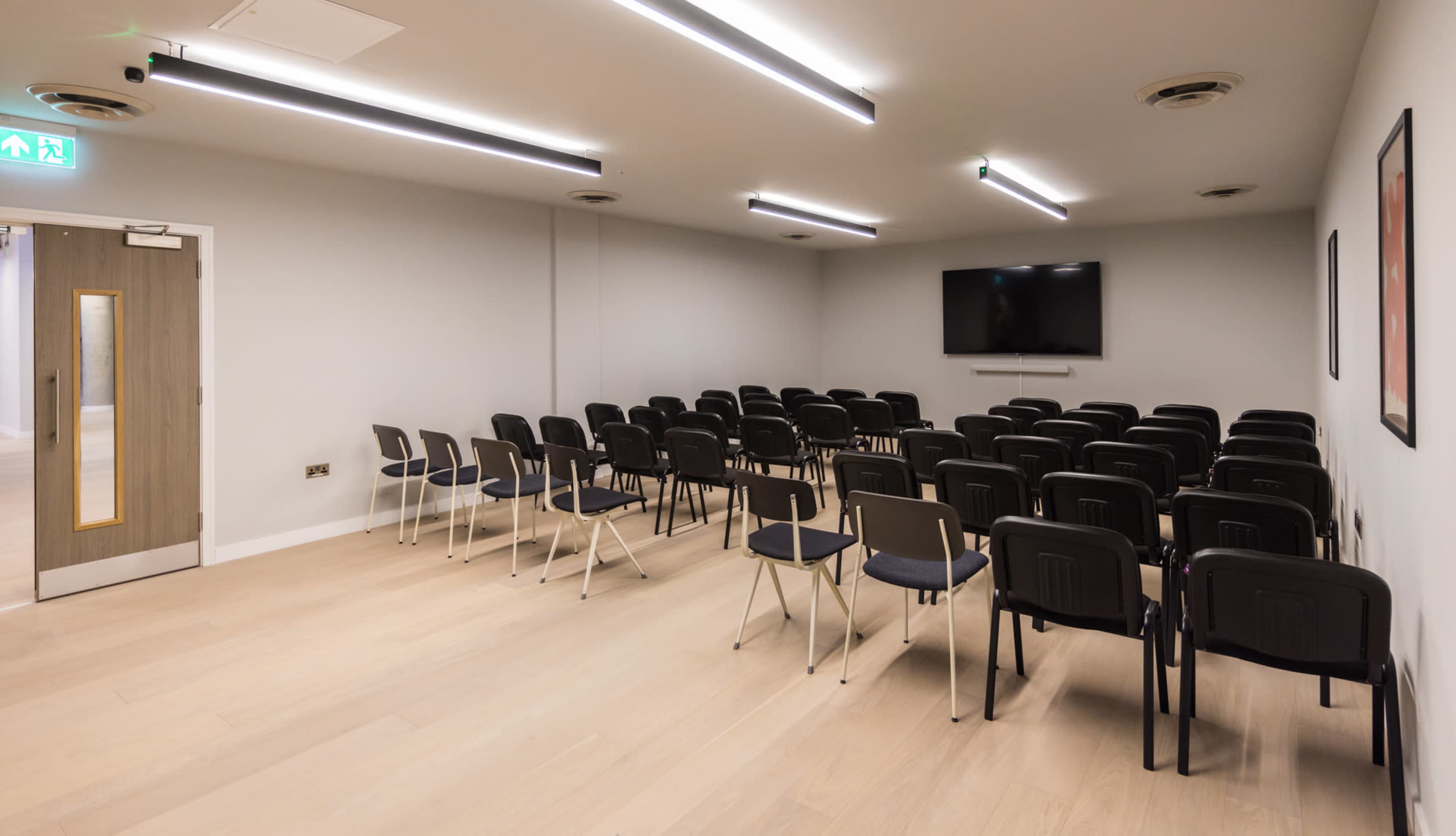 The image shows a meeting room with rows of black chairs facing a wall-mounted television.