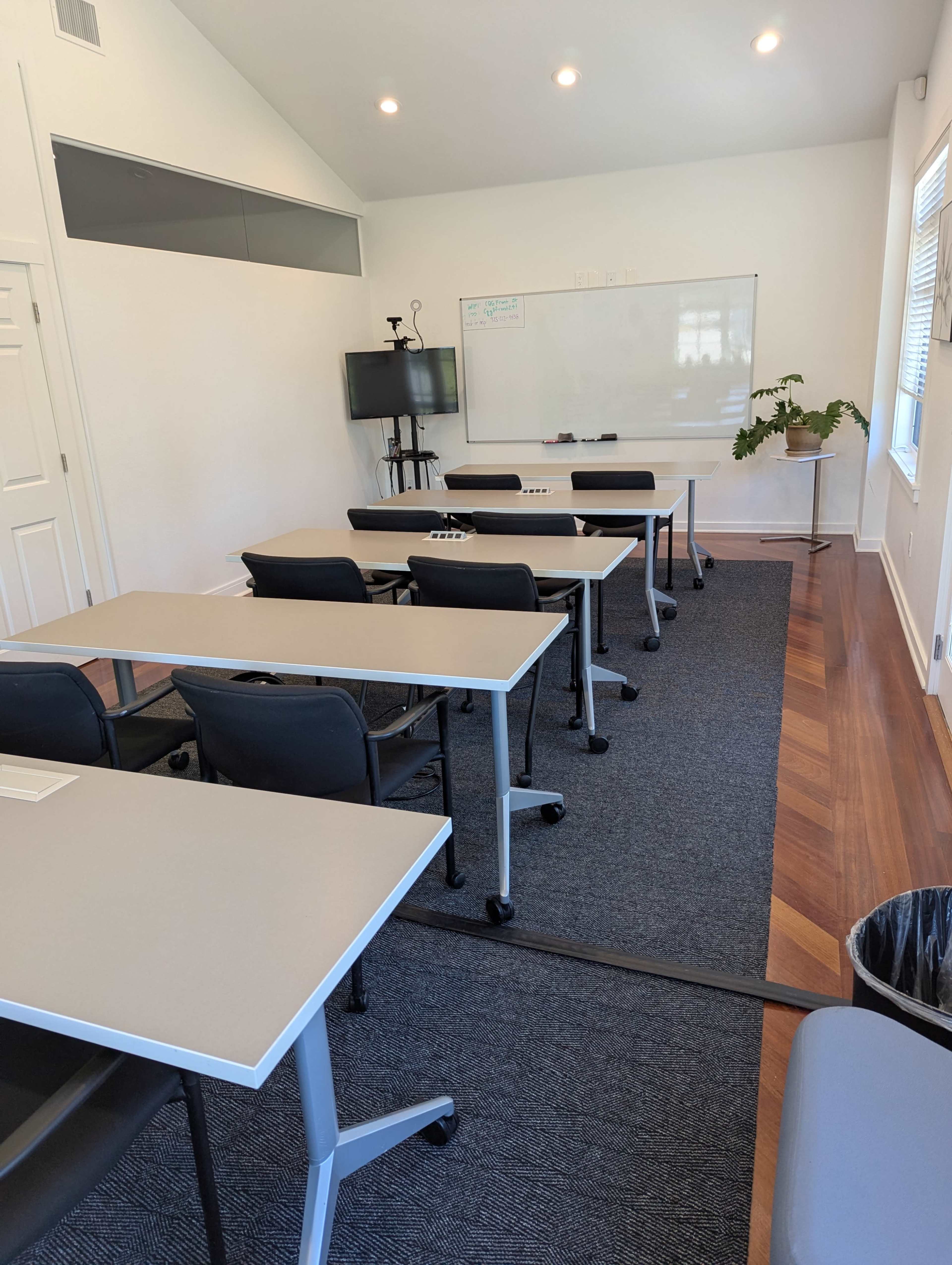 The image shows a classroom setting with several rows of desks, a whiteboard at the front, and a television mounted on the wall.