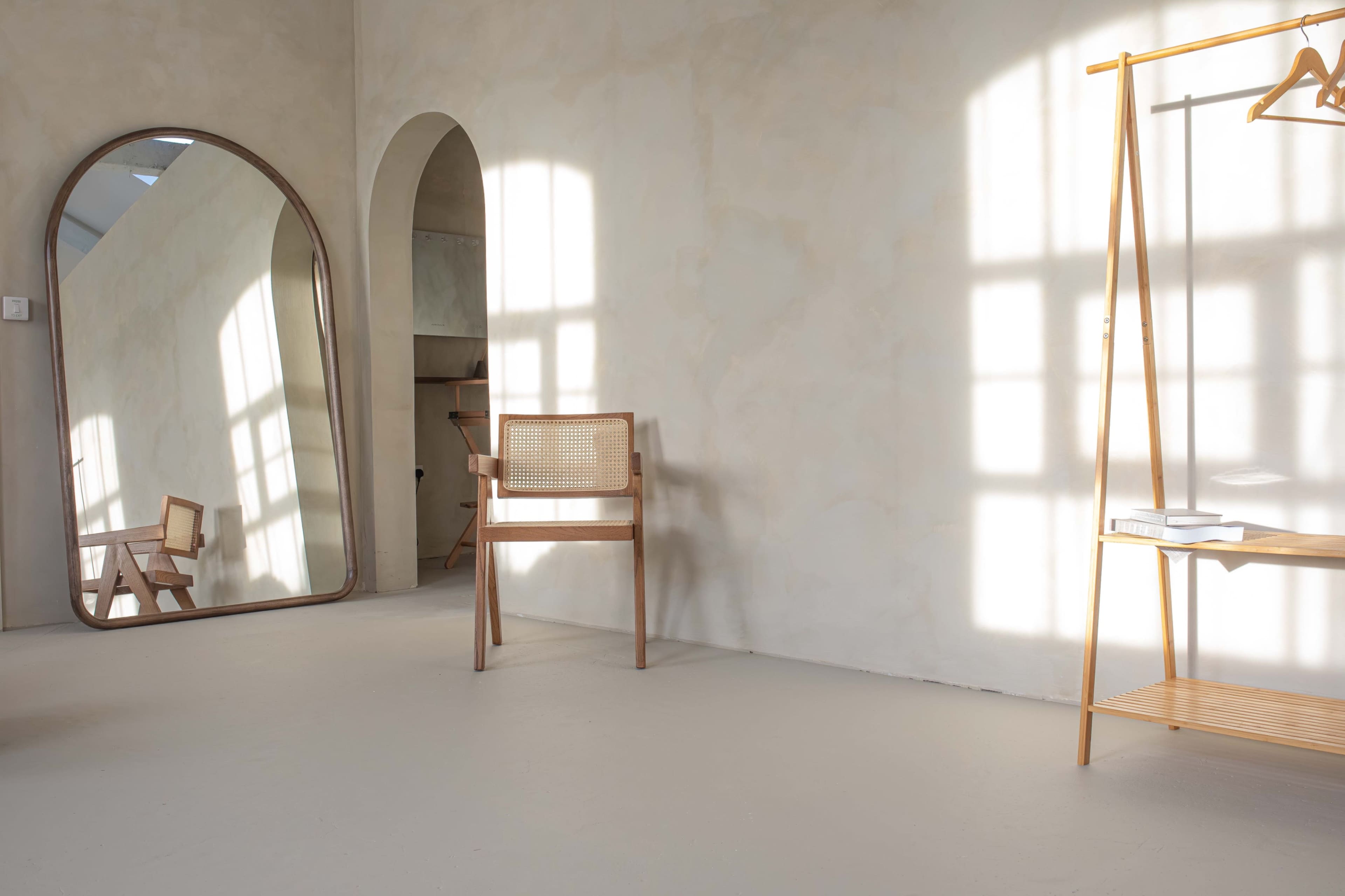 The image depicts a minimalist room with a large mirror, a wooden chair, and a minimalist clothing rack, all illuminated by soft sunlight filtering through the windows.