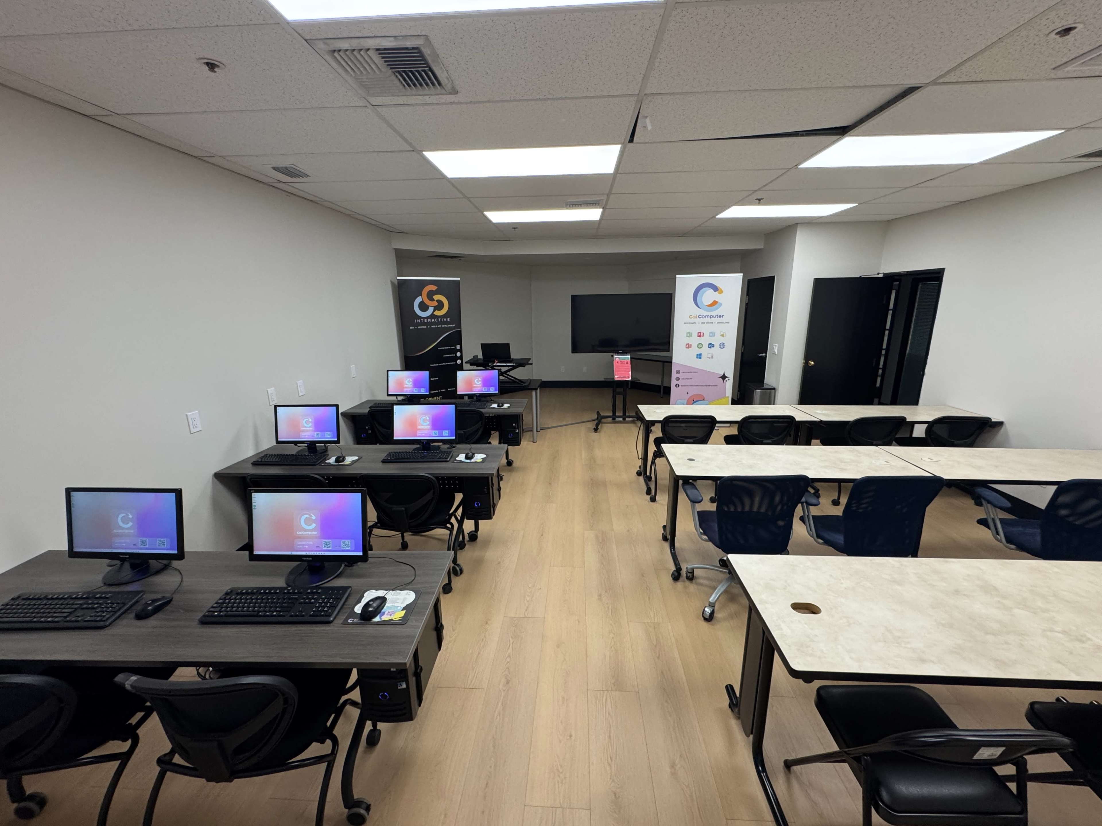 The image shows a classroom or training room with several computer desks arranged in rows, a large screen at the front, and banners on display.